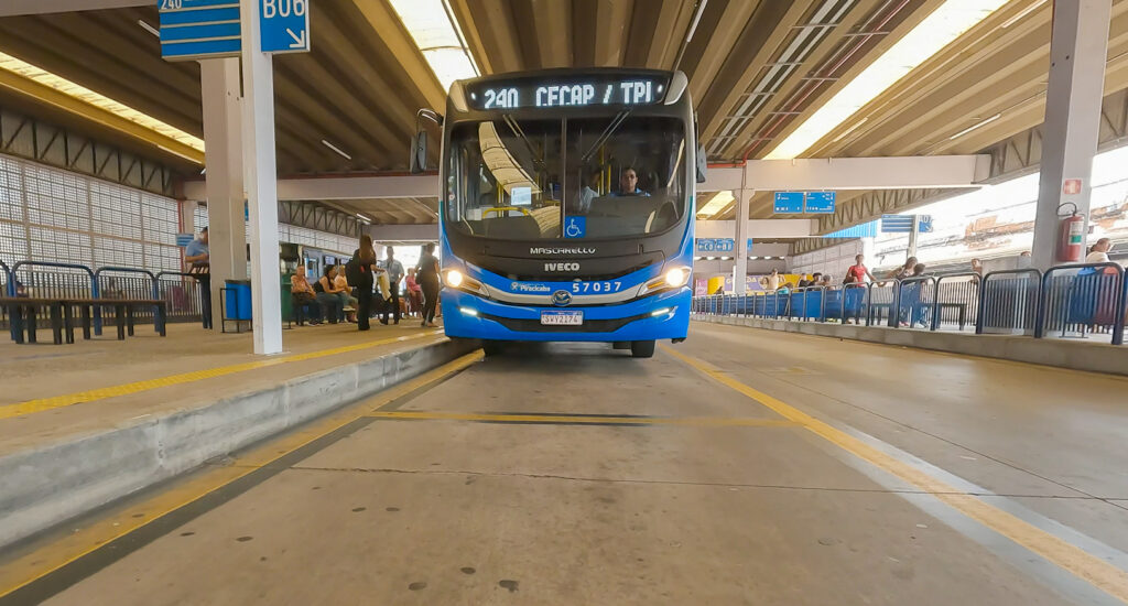 A imagem mostra um ônibus azul e branco da marca Mascarello, modelo Iveco, estacionado em um terminal de ônibus. O ônibus está posicionado entre plataformas, com uma faixa amarela de segurança no chão. No letreiro digital frontal do ônibus está escrito "240 CECAP / TPI", indicando a linha ou destino. Há pessoas aguardando na plataforma à esquerda, algumas sentadas e outras em pé. O terminal possui um teto de concreto com uma estrutura em relevo e iluminação natural que entra pelas laterais. O ambiente parece ser um local de embarque e desembarque de passageiros, possivelmente em Piracicaba, como sugere a inscrição na lateral do ônibus. A foto foi tirada em um ângulo que destaca a frente do veículo, com boa iluminação e foco nítido.