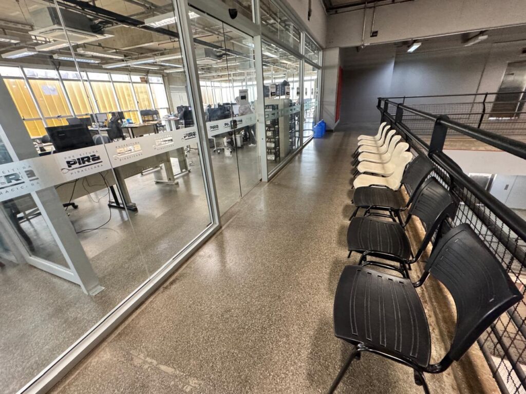 A imagem retrata um espaço de escritório moderno e um corredor adjacente, transmitindo uma atmosfera de funcionalidade e organização. Em detalhe, a imagem é dividida em duas áreas principais. À esquerda, através de uma parede de vidro transparente com moldura branca, observa-se um escritório amplo e moderno. O vidro reflete ligeiramente o ambiente externo, mas permite ver o interior, onde se encontram múltiplos postos de trabalho equipados com computadores e cadeiras de escritório. Há pessoas sentadas em algumas mesas, trabalhando. No vidro, há logotipos ou inscrições em branco que parecem ser "PREFEITURA" e "PIRA", possivelmente de "PIRACICABA", indicando que pode ser um ambiente administrativo ou público. O chão do escritório é de um material claro e liso. As janelas do escritório, visíveis ao fundo, revelam uma estrutura amarela no exterior, sugerindo a presença de um edifício próximo ou uma proteção solar. Do lado direito da imagem, e um pouco à frente do plano do vidro, há um corredor ou área de espera com um chão de cimento polido e granulado, que se estende por toda a largura da imagem. Ao longo de uma grade escura de metal, que serve como corrimão ou proteção de um nível inferior, estão alinhadas várias cadeiras. Grande parte dessas cadeiras é preta, de plástico e com estrutura de metal, mas algumas, no meio da fila, são de cor creme ou bege, contrastando com as pretas. A grade escura é robusta e tem um padrão de malha. Mais ao fundo do corredor, há uma parede cinza e um portão azul ou objeto azul vibrante que se destaca. O teto do corredor é de concreto exposto, com encanamentos e instalações elétricas visíveis, o que confere um toque industrial ao design do espaço. A iluminação parece ser artificial, com luminárias no teto. Não há pessoas visíveis no corredor, e a disposição das cadeiras sugere uma área de espera. A atmosfera geral é de um ambiente profissional e limpo. A luz dispersa, provavelmente diurna através das janelas combinada com iluminação artificial, cria um ambiente claro, mas sem luz solar direta forte, contribuindo para uma sensação de tranquilidade e seriedade no local de trabalho.