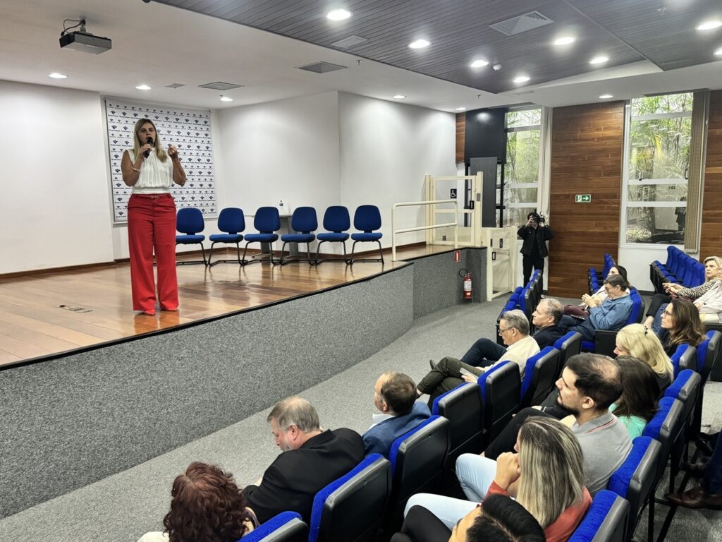 A imagem mostra uma palestra ou apresentação acontecendo em um auditório moderno. No centro do palco de madeira, uma mulher loira está falando ao microfone. Ela veste uma blusa branca sem mangas e calças largas vermelhas. Atrás dela, há seis cadeiras azuis vazias alinhadas e um painel de fundo com um logotipo repetido, indicando que o evento é formal ou corporativo. À direita do palco, há um elevador de acessibilidade e uma porta com uma placa de saída de emergência iluminada. O público, composto por cerca de 15 pessoas, está sentado em cadeiras acolchoadas azuis, prestando atenção à palestrante. A maioria dos espectadores usa roupas casuais de trabalho. O ambiente é bem iluminado, com luzes embutidas no teto escuro e grandes janelas que permitem a entrada de luz natural e mostram árvores do lado de fora. Há também um projetor instalado no teto e um extintor de incêndio próximo ao palco. A atmosfera geral é profissional e educacional, adequada para uma conferência, seminário ou treinamento.