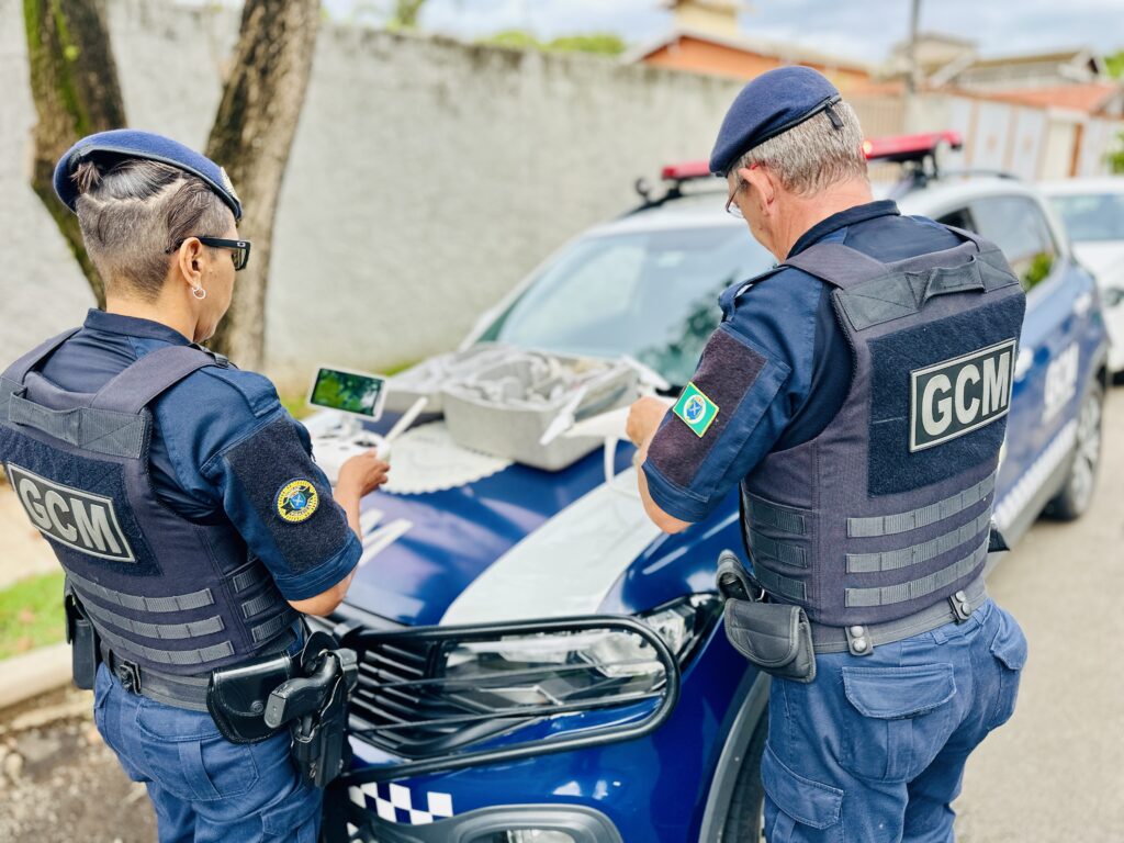 A imagem mostra dois agentes da Guarda Civil Municipal (GCM) em uma rua, ao lado de uma viatura policial azul e branca. A agente à esquerda é uma mulher, vestindo uniforme azul escuro com colete tático onde está escrito "GCM" nas costas, e um boina azul. Ela está segurando o controle remoto de um drone, com uma pequena tela visível. O agente à direita é um homem mais velho, também com uniforme e colete tático da GCM, olhando para um drone branco que está apoiado no capô da viatura. Ambos os agentes têm armas de fogo presas ao cinto. A viatura tem uma faixa xadrez na dianteira e uma barra de luz no teto. O ambiente é externo, com uma parede de concreto e algumas plantas ao fundo, sob luz natural do dia. Os uniformes têm distintivos, incluindo a bandeira do Brasil. A foto está com foco nos agentes e no drone, com o fundo levemente desfocado.