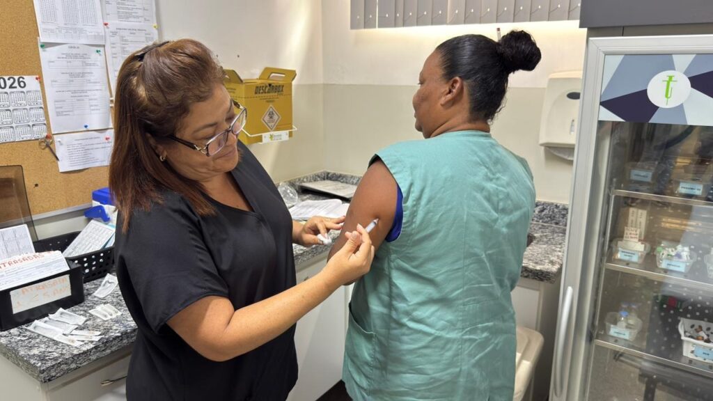 A imagem mostra um ambiente clínico onde uma profissional de saúde está aplicando uma vacina em uma paciente. A profissional, com cabelos castanhos e usando óculos e uniforme preto, está concentrada enquanto injeta a vacina no braço esquerdo da paciente, que veste um colete verde e tem o cabelo preso em um coque. Ao redor, há diversos elementos típicos de um consultório ou sala de vacinação, como uma caixa para descarte de materiais perfurocortantes (com a palavra "DESCARBOX"), um quadro de avisos com papéis afixados e uma geladeira específica para armazenamento de vacinas. No quadro, há inscrições como "ATRASADAS", "ATRASADAS < 5 ANOS" e "PNEUMO 10", indicando controle e organização das vacinas. O ambiente é bem iluminado e transmite uma sensação de cuidado e profissionalismo na aplicação das vacinas.