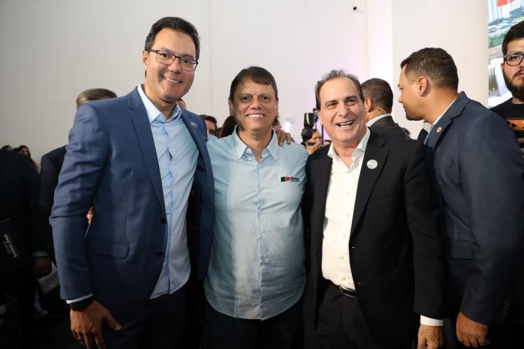 A imagem mostra quatro homens posando para uma foto em um ambiente interno, possivelmente em um evento profissional ou social. Três deles estão em primeiro plano, sorrindo e olhando diretamente para a câmera. O homem à esquerda veste um paletó azul e uma camisa social clara, usando óculos. O homem no centro está com uma camisa azul clara com um logotipo no peito e tem o braço apoiado no ombro do homem à esquerda. O homem à direita usa um paletó preto e camisa social branca, com um broche preso na lapela do paletó. O quarto homem está parcialmente visível à direita, vestindo um paletó azul escuro. O fundo é simples, com uma parede branca e alguns elementos pouco definidos, sugerindo um ambiente moderno e bem iluminado. No canto inferior da imagem, aparecem os nomes "MADALENA", "RICARDO" e "SÃO PAULO", possivelmente indicando nomes das pessoas ou locais relacionados à foto.