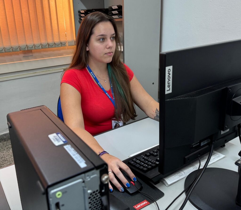 A imagem mostra uma mulher jovem sentada em uma mesa de escritório, trabalhando em um computador Lenovo. Ela tem cabelos longos castanhos com mechas azul-esverdeadas e está usando uma camiseta vermelha e um crachá azul com a palavra "semae" repetida. A mulher está concentrada, olhando para o monitor à sua frente, com a mão direita no mouse. Suas unhas estão pintadas de azul com glitter. No braço esquerdo, ela tem uma pequena tatuagem de borboleta. A mesa é branca e há um teclado preto à sua frente. Ao fundo, há uma janela com persianas verticais laranja e uma estante com pastas organizadas. A iluminação é clara e a foto utiliza uma profundidade de campo que mantém a mulher em foco, enquanto o computador em primeiro plano está levemente desfocado.