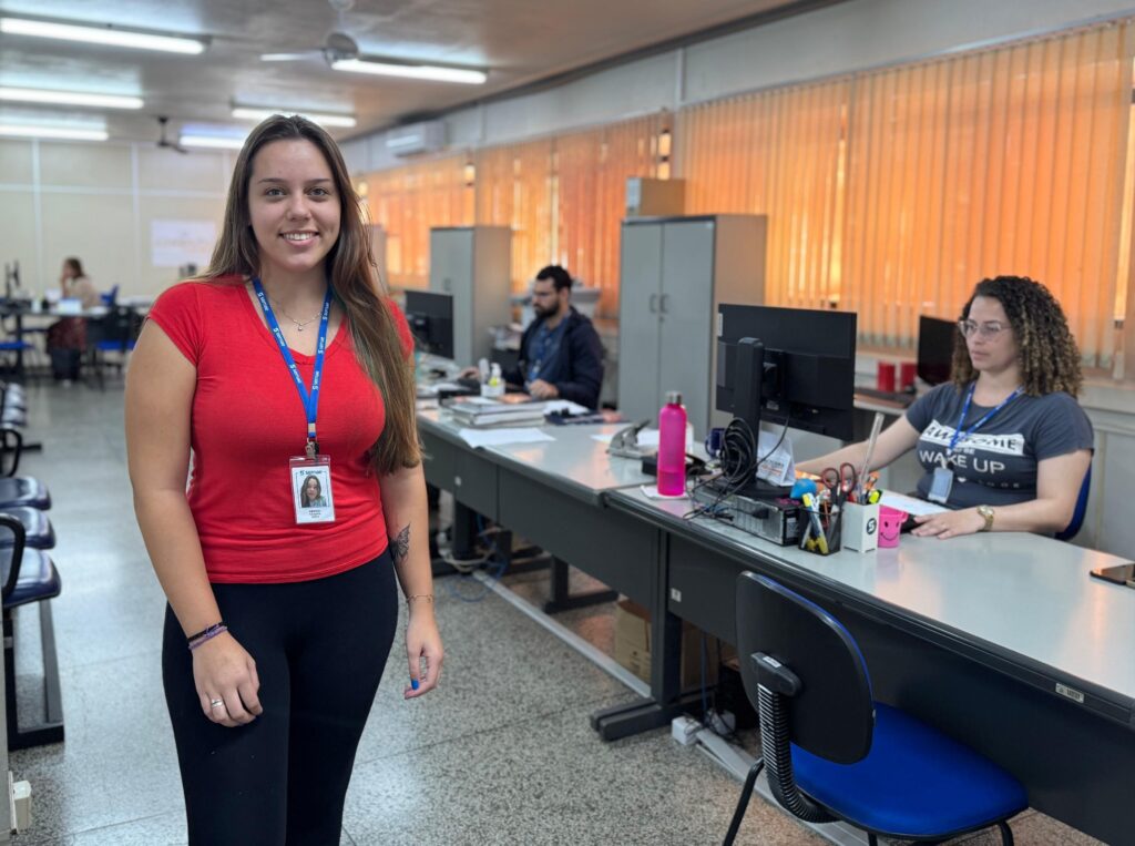 A imagem mostra uma mulher sorridente em primeiro plano, vestindo uma camiseta vermelha e calça preta. Ela usa um crachá azul pendurado no pescoço, que exibe sua foto e o nome "Amanda". No braço esquerdo, ela tem uma tatuagem de uma borboleta. Ao fundo, há um ambiente de escritório com várias mesas e computadores, onde duas outras pessoas estão trabalhando. Uma delas é uma mulher vestindo uma camiseta preta com as palavras "AWSOME WAKE UP" impressas. O escritório possui iluminação fluorescente e janelas com persianas verticais. O ambiente parece ser um local de trabalho típico, organizado e bem iluminado. A palavra "semae" aparece repetidamente nos crachás, sugerindo o nome da empresa ou organização.
