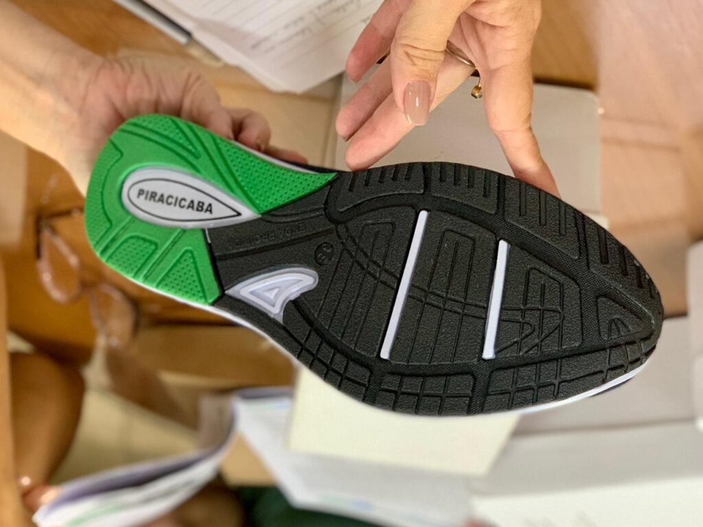 A imagem mostra as mãos de uma pessoa segurando um tênis de forma que a sola fique voltada para a câmera. A sola do tênis é predominantemente preta, com uma parte verde vibrante no calcanhar. No centro da parte verde, há um logotipo branco oval com a palavra "PIRACICABA". Na sola preta, há um padrão complexo de sulcos e texturas para aderência, além de duas barras brancas verticais próximas à parte frontal do pé. Também é possível ver o número "38", que indica o tamanho do calçado, e a inscrição "VENDA PROIBIDA" gravada na sola. As mãos da pessoa que segura o tênis têm pele clara; a mão direita tem unhas feitas com esmalte nude e um anel dourado no dedo anelar. O fundo é desfocado, mas é possível identificar uma superfície de madeira e algumas folhas de papel, sugerindo um ambiente interno, como uma loja ou escritório. A iluminação é clara, destacando os detalhes da sola do tênis.