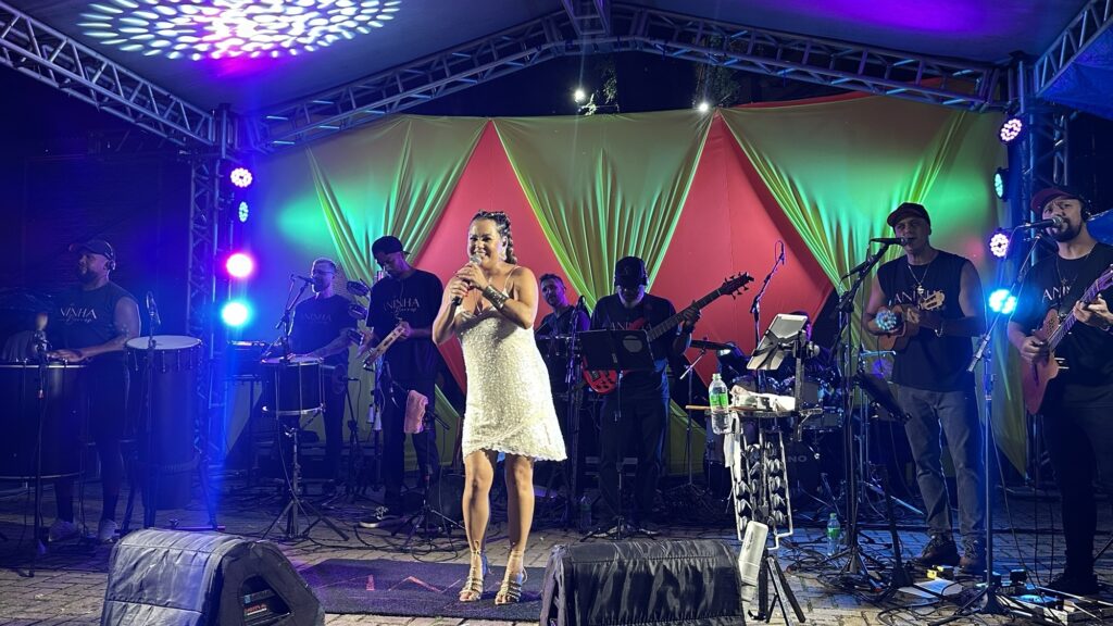 A imagem mostra uma apresentação musical ao vivo em um palco ao ar livre durante a noite. No centro, há uma cantora mulher vestindo um vestido branco com brilho, segurando um microfone e sorrindo enquanto canta. Ela está rodeada por seis músicos, todos homens, que tocam diferentes instrumentos, como violão, cavaquinho, baixo, tamborim e pandeiro. A maioria dos músicos veste camisetas pretas com a inscrição "ANINHA Barros", que provavelmente é o nome do grupo ou da cantora. O palco está decorado com panos coloridos em tons de amarelo e vermelho ao fundo, e a iluminação é vibrante, com luzes roxas, azuis e rosas que criam um ambiente festivo e animado. O cenário sugere uma apresentação de samba ou pagode, gêneros musicais típicos do Brasil. A disposição dos músicos é organizada, com a cantora à frente e os instrumentistas distribuídos ao redor dela, compondo uma atmosfera de interação e energia musical.