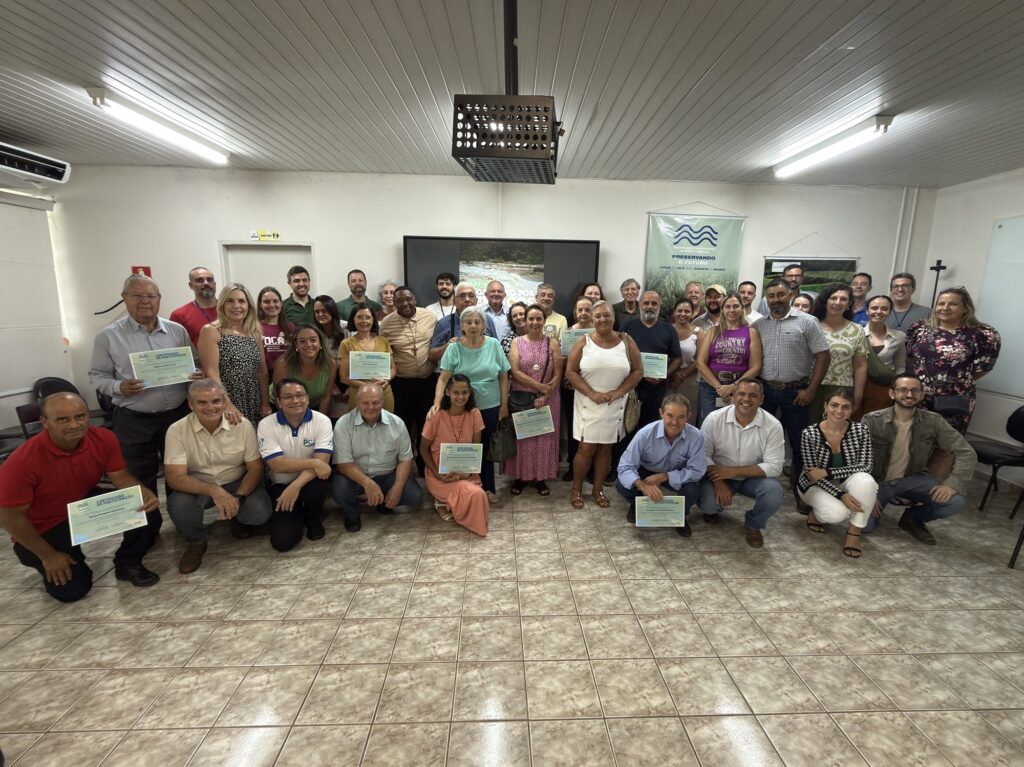 A imagem mostra um grande grupo de pessoas reunidas para uma foto em um ambiente interno, possivelmente uma sala de eventos ou uma sala de reuniões. São cerca de 45 pessoas de diferentes idades e gêneros, muitas delas segurando certificados de participação, indicando que provavelmente participaram de um curso, workshop ou programa. Ao fundo, há uma tela grande com uma imagem que parece ser de um rio, e um banner com os dizeres "PRESERVANDO O FUTURO" e "CUIDAR DO HOJE PARA GARANTIR O AMANHÃ". Também é possível ver que o evento está relacionado a um "PROGRAMA MUNICIPAL DE PAGAMENTO POR SERVIÇOS AMBIENTAIS", sugerindo que o grupo está envolvido em atividades ambientais ou de preservação. As pessoas estão organizadas em várias filas, algumas em pé e outras agachadas na frente, todas olhando para a câmera e sorrindo, demonstrando um clima de celebração e conquista. O ambiente é bem iluminado, com teto branco e luminárias fluorescentes, além de um piso claro com padrão marmorizado. O conjunto passa uma sensação de união e compromisso com a causa ambiental.