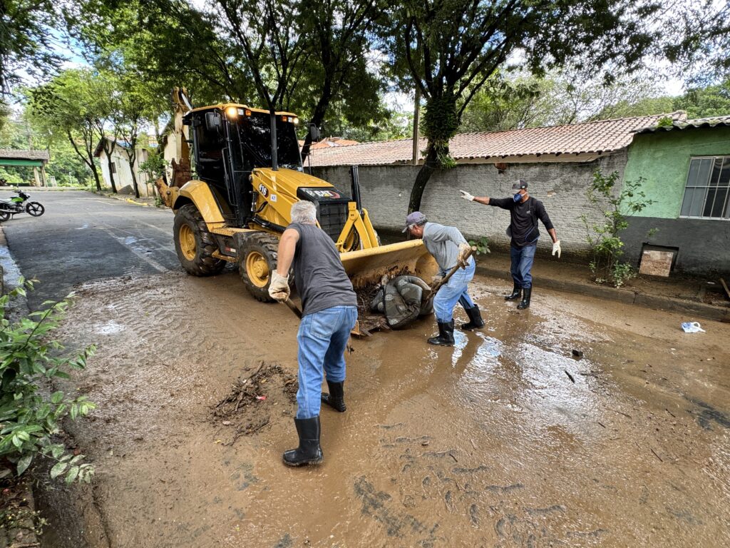 A imagem mostra uma cena de limpeza em uma rua residencial coberta de lama. Três homens estão trabalhando para remover a sujeira e detritos do local. Um deles, mais próximo à câmera, segura uma pá e está de costas, usando uma camiseta cinza, calça jeans e botas pretas de borracha. Outro homem, ao lado da máquina, também está usando uma pá para empurrar a lama para dentro da pá da retroescavadeira amarela da marca CAT, modelo 416. Um terceiro homem, vestindo camiseta preta, calça jeans e máscara no rosto, aponta para algo à frente. Ao fundo, casas simples com telhados de cerâmica e paredes pintadas em tons claros, além de árvores verdes que cercam a rua. O céu está nublado, indicando um dia possivelmente chuvoso ou recente. A cena sugere um trabalho de limpeza após um evento de chuva forte ou enchente.