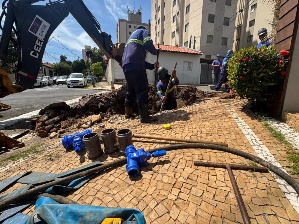 A imagem mostra uma cena de construção em uma calçada de cidade durante o dia. No primeiro plano, há vários materiais de encanamento, como válvulas azuis, segmentos de tubos cinzas, uma mangueira longa e uma ferramenta metálica em formato de T, todos sobre um piso de paralelepípedos. No centro da imagem, dois trabalhadores vestindo uniformes azuis com faixas refletivas amarelas estão trabalhando em uma vala aberta no chão. Um deles está em pé na borda da vala usando uma barra metálica longa, enquanto o outro está parcialmente dentro da vala. À esquerda, o braço preto de uma escavadeira com a marca "DEERE" e o logo "semae" está visível. Ao fundo, é possível ver prédios residenciais de vários andares, carros estacionados na rua e outros trabalhadores próximos. A luz do sol é forte, criando sombras nítidas, e a imagem transmite a ideia de um projeto urbano de manutenção, combinando trabalho manual e uso de máquinas.
