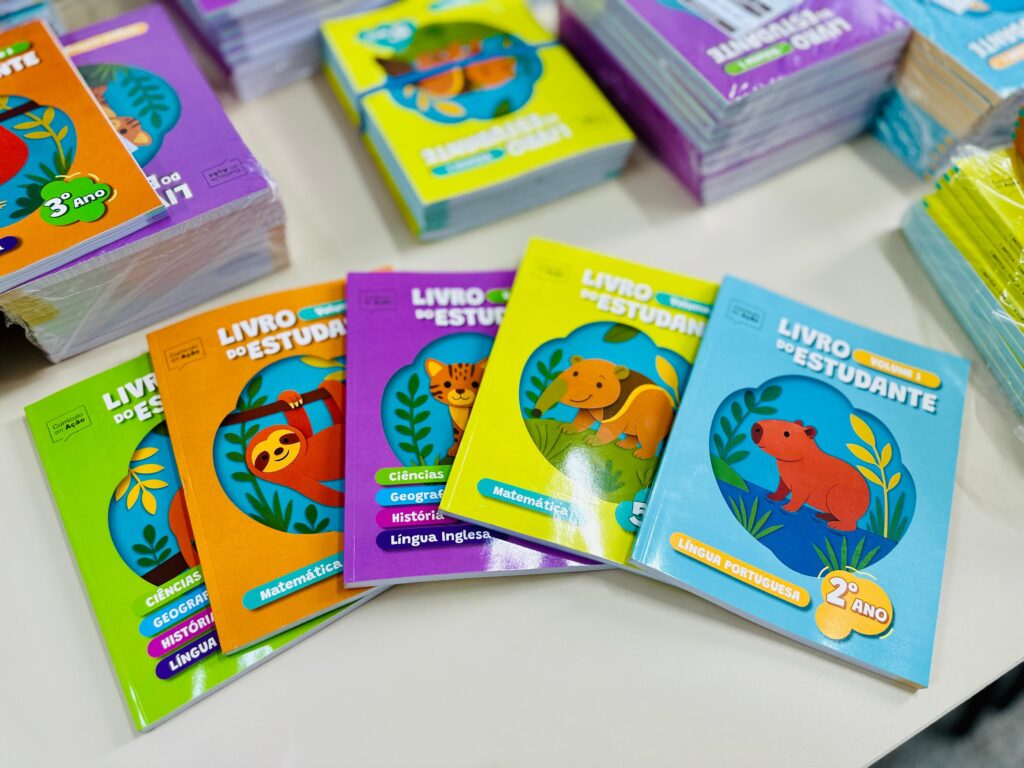 A imagem mostra uma coleção de livros didáticos intitulados "LIVRO DO ESTUDANTE", voltados para o ensino fundamental, especificamente para o 2º e 3º anos. Os livros possuem capas coloridas e vibrantes, cada uma com uma cor diferente (verde, laranja, roxa, amarela e azul claro) e uma ilustração circular de um animal na parte frontal, como uma preguiça, um felino, um tamanduá e uma capivara. Os títulos indicam que os livros abrangem várias disciplinas escolares, incluindo Ciências, Geografia, História, Língua Inglesa, Matemática e Língua Portuguesa. O texto "Currículo em Ação" sugere que esses livros são parte de um programa educacional. Ao fundo, há várias pilhas desses mesmos livros, alguns ainda embalados em plástico, o que indica que são novos e possivelmente prontos para distribuição em escolas. A imagem está bem iluminada e foca nos livros dispostos em primeiro plano, com um leve desfoque nas pilhas ao fundo.