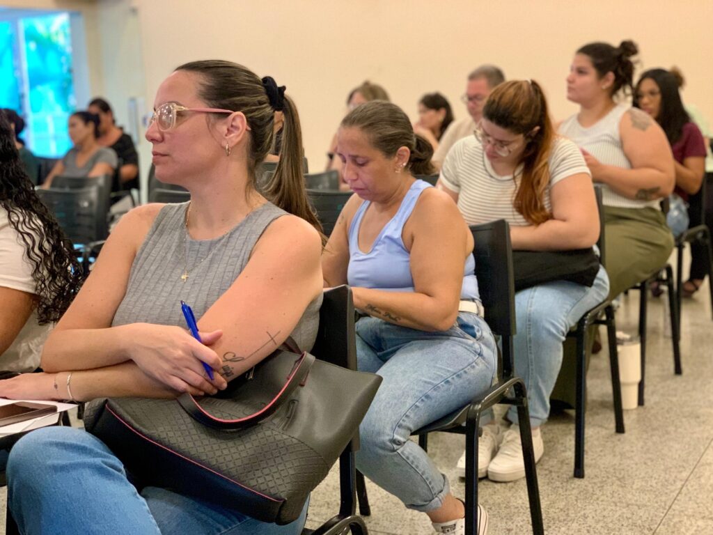 A imagem mostra um grupo de adultos sentados em cadeiras, provavelmente participando de uma aula, palestra ou workshop. O foco principal está em uma mulher na frente, que usa óculos, uma blusa cinza de manga longa e calça jeans azul, segurando uma caneta azul na mão. Ao lado dela, há outra mulher com blusa regata azul clara e calça jeans, que está olhando para baixo, talvez lendo ou escrevendo algo. Ao fundo, é possível ver outras pessoas também sentadas, algumas aparentando prestar atenção e outras tomando notas. As cadeiras são pretas com estrutura metálica, dispostas em fileiras. O ambiente parece ser uma sala interna, com paredes claras e piso cinza. A iluminação é suave e natural, possivelmente vinda de janelas grandes ao fundo. Há uma bolsa preta com detalhes em rosa apoiada em uma das cadeiras. A atmosfera sugere um ambiente de aprendizado ou reunião, com os participantes atentos e engajados no que está sendo apresentado.