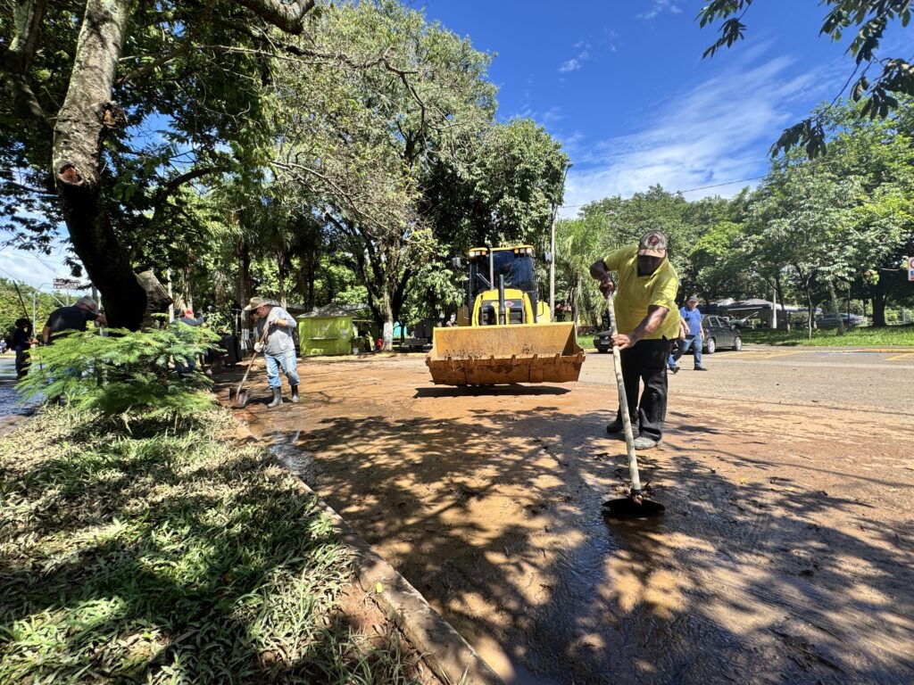 A imagem mostra um grupo de trabalhadores removendo uma grande quantidade de lama de uma estrada pavimentada, provavelmente após uma enchente ou deslizamento de terra. No centro da cena, há uma retroescavadeira amarela com a pá baixa, ajudando na limpeza. À frente, em destaque, um homem com camiseta amarela, calça preta e boné usa uma enxada para auxiliar na remoção da lama. Outros trabalhadores, vestidos com roupas casuais e botas de borracha, também estão usando pás e enxadas para limpar o local. A estrada está cercada por árvores verdes e o céu está claro, com poucas nuvens, indicando um dia ensolarado. Ao fundo, é possível ver uma pequena estrutura verde, possivelmente um quiosque ou ponto de comércio. A cena transmite um esforço coletivo para recuperar a via após um evento natural que causou o acúmulo de lama.