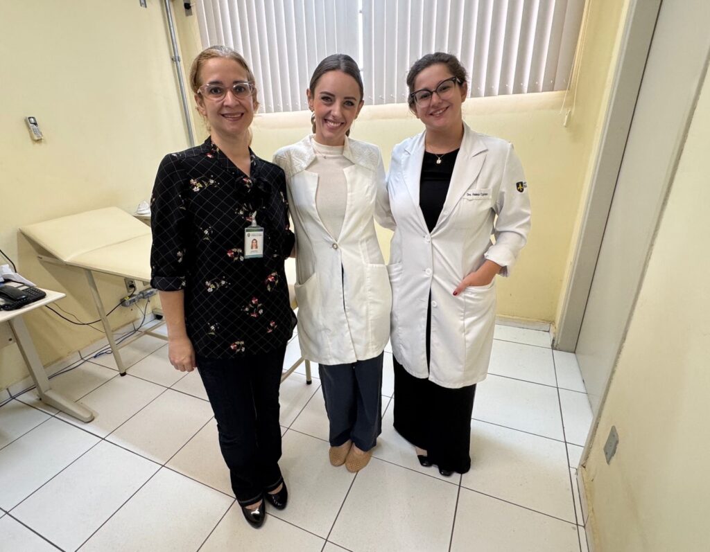 A imagem mostra três mulheres em um ambiente clínico, possivelmente um consultório médico. Elas estão em pé, lado a lado, sorrindo para a câmera. A mulher à esquerda veste uma blusa preta com estampa e calça escura, enquanto as outras duas usam jalecos brancos, indicando que são profissionais da área da saúde. A mulher no centro usa uma blusa de gola alta branca por baixo do jaleco, e a mulher à direita usa uma saia preta longa sob o jaleco, que tem um brasão na manga esquerda. No jaleco dela há um crachá com o nome "Dra. Helena Cypriano". O ambiente é bem iluminado, com paredes claras, uma mesa de exame médica à esquerda e uma pequena mesa com um telefone ao fundo, próximo a uma janela com persianas verticais. O cenário sugere um consultório ou clínica médica durante o dia.