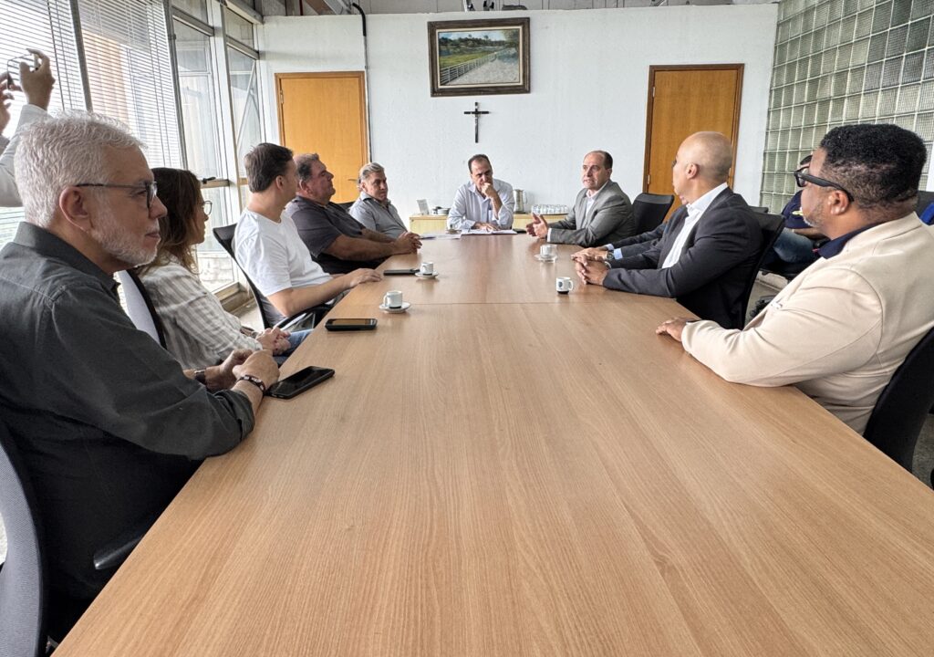 A imagem mostra uma reunião formal em um ambiente de escritório. Nove pessoas estão sentadas ao redor de uma mesa retangular longa de madeira clara. A mesa está disposta horizontalmente na imagem, com cinco pessoas do lado esquerdo, uma pessoa na cabeceira e três pessoas do lado direito. A pessoa na cabeceira parece liderar a reunião, vestindo uma camisa azul clara. À esquerda, há quatro homens e uma mulher, todos vestidos de forma casual ou semi-formal. À direita, três homens vestidos de forma mais formal, com blazers e ternos. Um dos homens à direita está fazendo um gesto com as mãos, como se estivesse explicando algo. Sobre a mesa, há vários copos pequenos de café e alguns celulares. O ambiente é claro, com janelas grandes à esquerda e paredes brancas. Na parede ao fundo, há uma pintura de paisagem e uma cruz preta fixa. Também há duas portas de madeira ao fundo e uma parede com blocos de vidro à direita. A atmosfera parece séria e profissional, indicando uma reunião de negócios ou política.