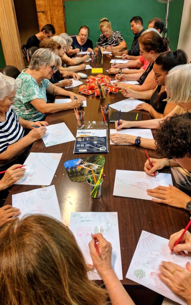 A imagem mostra um grupo grande de pessoas, principalmente idosos, sentados ao redor de uma longa mesa retangular de madeira escura, participando de uma oficina de desenho. Todos estão concentrados em suas folhas de papel, usando lápis de cor para criar seus desenhos. Na mesa, há vários copos plásticos cheios de lápis de cor, um pacote de papel com a marca "Report" e alguns petiscos em embalagens vermelhas. Os desenhos visíveis incluem casas, árvores e formas abstratas, mostrando que os participantes estão explorando temas variados. O ambiente parece ser uma sala interna bem iluminada, com paredes verdes ao fundo e uma porta de madeira à esquerda. A foto foi tirada de um ângulo elevado, permitindo ver toda a extensão da mesa e as pessoas envolvidas na atividade. A atmosfera parece tranquila e colaborativa, com todos focados na atividade artística, alguns até sorrindo, sugerindo um momento agradável e de interação social.