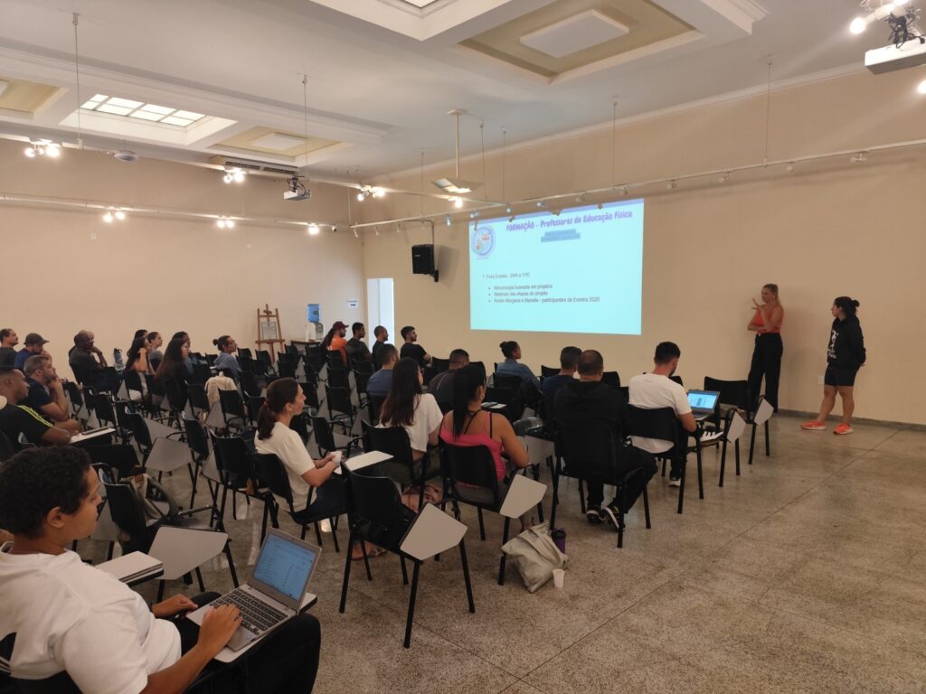 A imagem mostra uma sala de treinamento ou palestra, com várias pessoas sentadas em cadeiras com pequenas mesas acopladas, assistindo a uma apresentação projetada na parede. A apresentação, em português, tem o título "FORMAÇÃO – Professores de Educação Física" e lista alguns tópicos relacionados a um evento chamado "Feira Eureka" que ocorrerá entre 29 de setembro e 1º de outubro. Duas mulheres estão em pé próximas à projeção, uma delas parece estar apresentando enquanto a outra observa. A maioria dos participantes está atenta, alguns com notebooks abertos, sugerindo um ambiente de aprendizado ou capacitação. A sala é ampla, bem iluminada, com teto alto e iluminação embutida, e o piso é de um material brilhante, possivelmente granito ou porcelanato. Ao fundo, há um bebedouro e um cavalete de madeira. O ambiente transmite uma atmosfera profissional e colaborativa.