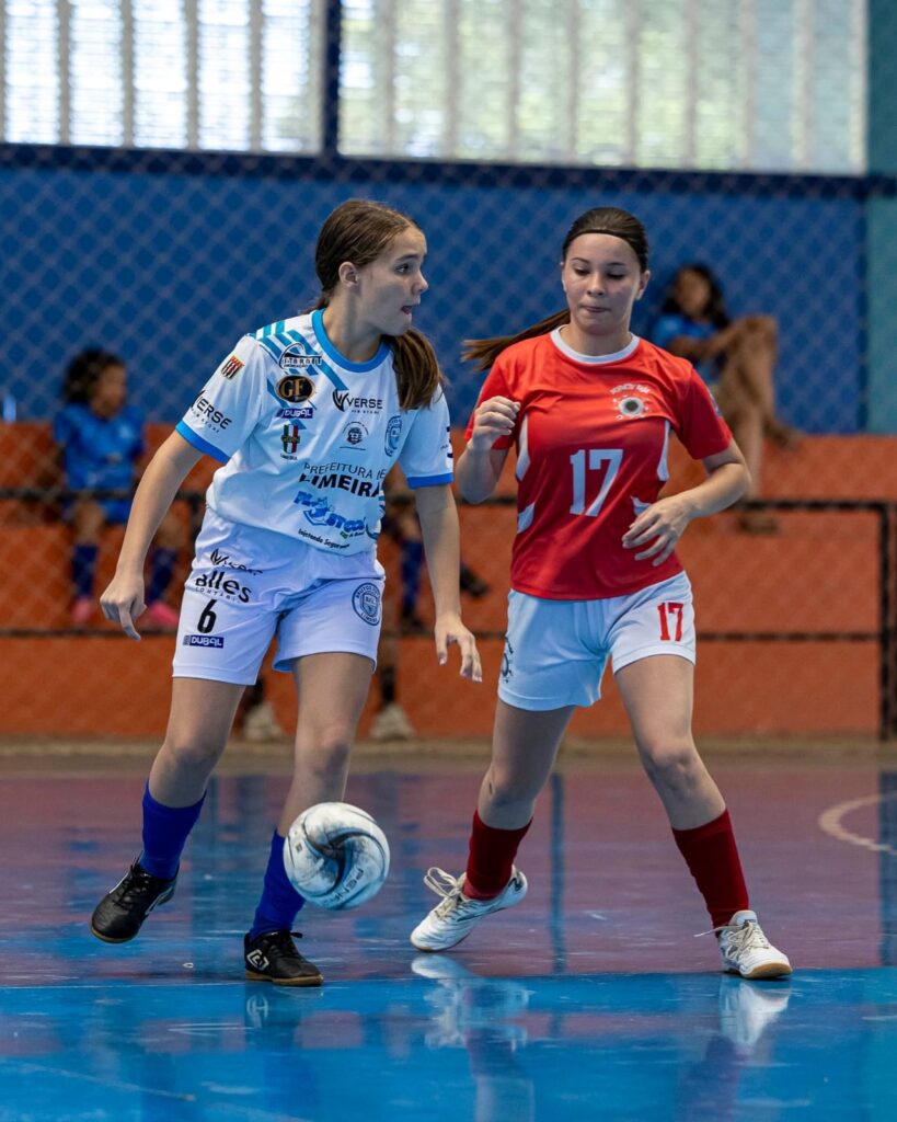 A imagem mostra uma cena de um jogo de futsal entre duas jogadoras femininas jovens. A jogadora à esquerda veste um uniforme branco com detalhes em azul e o número 6, enquanto controla a bola branca com detalhes azuis. Ela parece concentrada e está avançando com a bola. A jogadora à direita usa um uniforme vermelho com o número 17 e está em uma posição defensiva, tentando bloquear a adversária. O jogo ocorre em uma quadra azul dentro de um ginásio, com arquibancadas laranja e uma grade azul ao fundo. Algumas pessoas estão assistindo à partida nas arquibancadas. A iluminação é clara, destacando a ação das atletas. O ambiente sugere uma competição organizada, com patrocinadores visíveis nos uniformes das jogadoras.