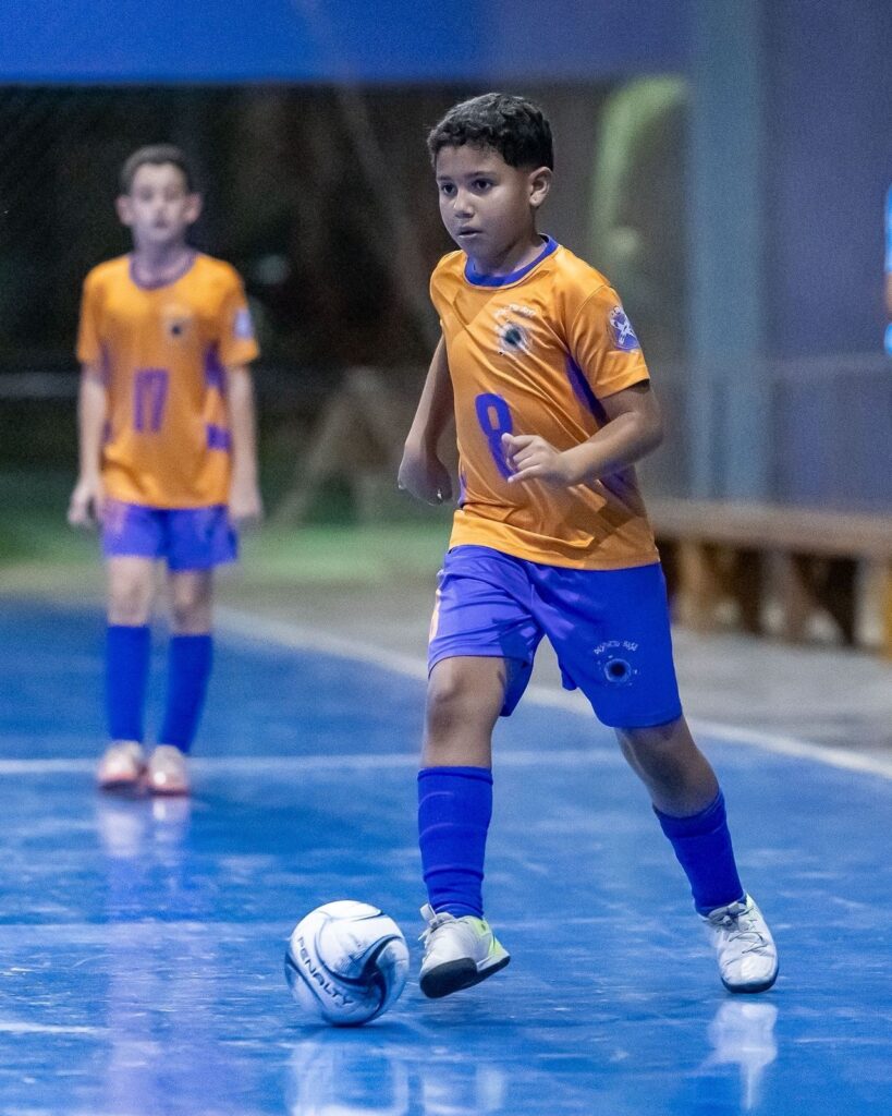 A imagem mostra um garoto jogando futsal em uma quadra azul coberta. Ele está em destaque, vestindo uma camisa laranja com detalhes roxos e o número 8, além de shorts roxos e meias azuis. O menino está em movimento, controlando a bola branca com detalhes pretos com o pé esquerdo, e parece concentrado no jogo. Ao fundo, desfocado, há outro garoto também com uniforme laranja com o número 17, observando a cena. O ambiente é iluminado, e ao lado direito há um banco de madeira encostado na parede cinza. A foto transmite a energia e o foco de uma partida de futsal entre jovens.