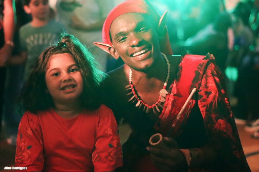 A imagem mostra uma cena com duas pessoas em destaque: uma menina jovem e um homem vestido como o Saci-Pererê, personagem folclórico brasileiro. A menina está à esquerda, sorrindo, com cabelos escuros e cacheados presos em um pequeno rabo de cavalo no topo da cabeça. Ela veste uma blusa vermelha com detalhes bordados nas mangas. Ao lado direito, o homem fantasiado de Saci-Pererê também sorri para a câmera. Ele usa um gorro vermelho característico do personagem, orelhas pontudas falsas, um colar de contas brancas com pendentes marrons e uma faixa vermelha com padrões ao redor da cintura. Ele segura um cachimbo de madeira na mão esquerda. A iluminação da foto é dramática, com luz verde vindo da direita que cria um contraste marcante com os tons vermelhos das roupas. Ao fundo, desfocados, aparecem outras pessoas, sugerindo um ambiente festivo ou cultural. A imagem transmite alegria e celebração da cultura brasileira. No canto inferior esquerdo, há o nome "Aline Rodrigues", provavelmente o crédito da fotografia.