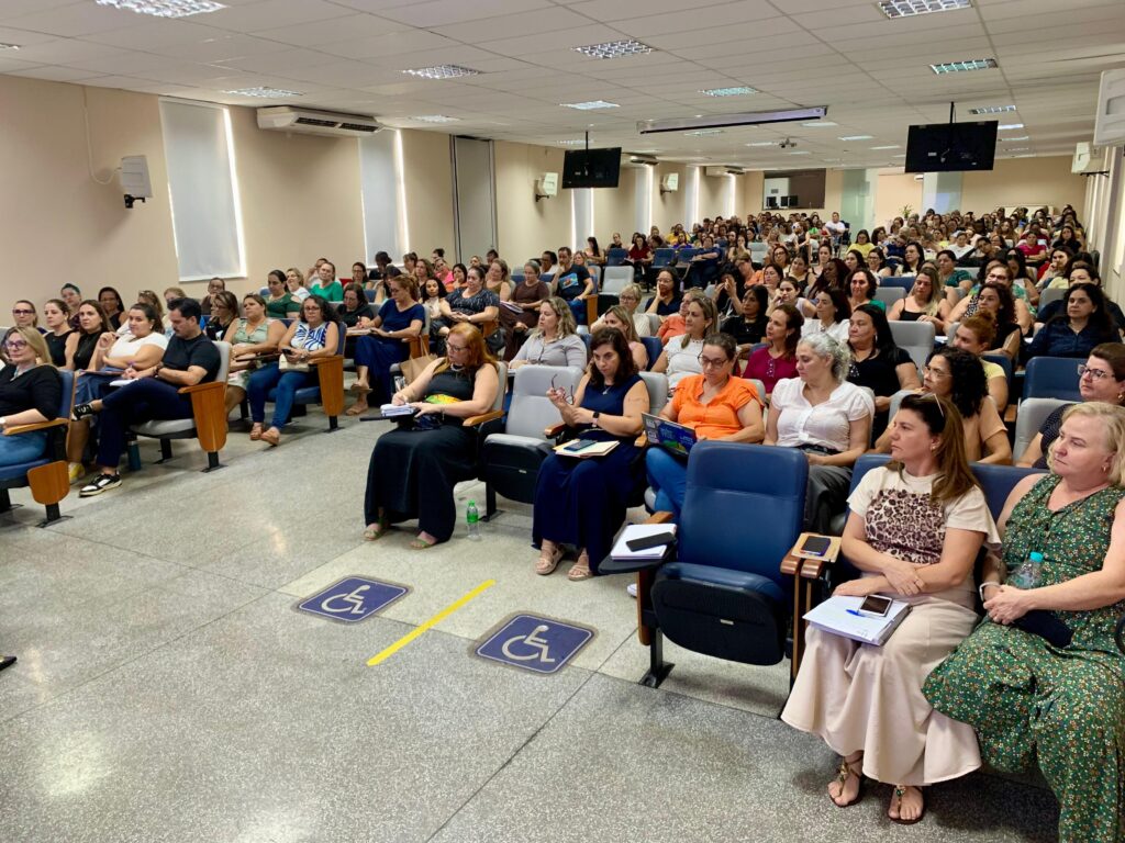 A imagem mostra um auditório ou sala de palestras cheia de pessoas sentadas em fileiras de cadeiras acolchoadas, organizadas em níveis. O ambiente é bem iluminado por luzes no teto e transmite uma atmosfera de aprendizado ou desenvolvimento profissional, já que muitos participantes estão com cadernos ou anotações em mãos. Na parte frontal, há dois espaços reservados para acessibilidade, demarcados com símbolos de cadeira de rodas e linhas amarelas no chão. As paredes brancas, os aparelhos de ar-condicionado e as grandes telas suspensas completam o cenário, reforçando a ideia de um espaço moderno e preparado para apresentações ou aulas coletivas.
