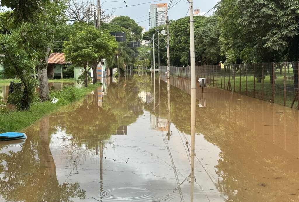 A imagem mostra uma rua residencial completamente alagada por água barrenta. A água cobre toda a extensão da via, submersa até quase o nível dos postes de luz que ficam ao longo da rua. De um lado, há uma cerca de arame parcialmente submersa e do outro, árvores verdes e arbustos que margeiam a rua. Ao fundo, é possível ver um prédio alto e moderno, indicando que a área é urbana ou suburbana. O céu está nublado, sugerindo que a chuva pode ter causado o alagamento recente. Não há pessoas visíveis na cena, apenas a água que domina o ambiente, refletindo os postes e as árvores. A sensação geral é de um cenário de enchente que interrompe a circulação e causa transtornos para os moradores locais.