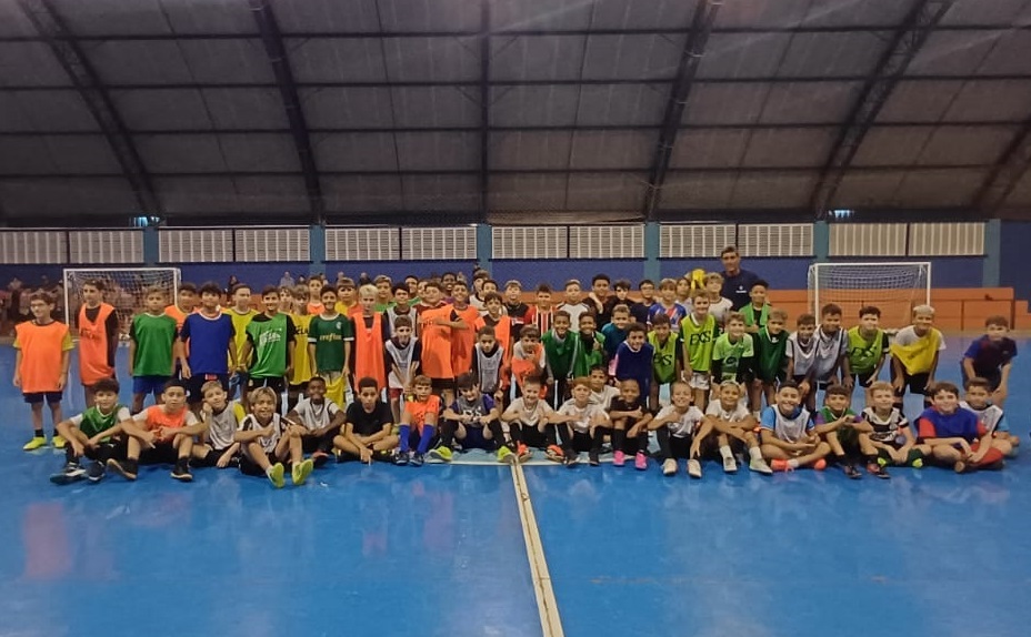 A imagem mostra um grande grupo de meninos, provavelmente entre 7 e 12 anos, reunidos para uma foto em uma quadra de futsal ou futebol de salão coberta e com piso azul. Eles estão organizados em várias fileiras, alguns agachados na frente e outros em pé atrás, todos vestidos com roupas esportivas coloridas, incluindo camisetas, coletes e uniformes de diferentes cores, como verde, laranja, amarelo e azul. Algumas camisetas verdes possuem a logo do Palmeiras e o patrocinador "Crefisa", indicando que podem ser de times ou escolinhas relacionadas a esse clube. Ao fundo, é possível ver duas traves de futsal e a estrutura da quadra indoor, com teto alto e iluminação artificial. Um adulto, provavelmente um treinador, está posicionado à direita, atrás das crianças. A cena transmite um ambiente de treinamento ou evento esportivo para jovens atletas.