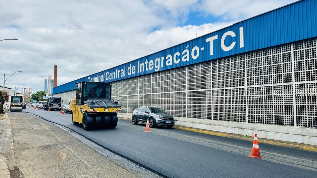 A imagem mostra uma obra de pavimentação em frente ao "Terminal Central de Integração - TCI", que é um grande terminal de ônibus com fachada azul e letras brancas. No primeiro plano, há uma máquina amarela da marca Caterpillar (CAT), um rolo compactador de pneus, que está operando sobre o asfalto recém-aplicado. Ao fundo, é possível ver outros veículos, incluindo um caminhão e outro rolo compactador, além de cones de sinalização laranja delimitando a área de trabalho. À direita, um carro cinza da marca Volkswagen está estacionado ou parado próximo ao meio-fio, separado da área de obra pelos cones. O céu está nublado, com algumas partes azuis visíveis, e ao fundo aparecem edifícios altos e uma chaminé industrial, indicando que o local é uma área urbana. A cena transmite a ideia de um trabalho de manutenção ou melhorias viárias em um importante ponto de integração de transporte público, com foco na pavimentação e compactação do asfalto para melhorar a infraestrutura local.