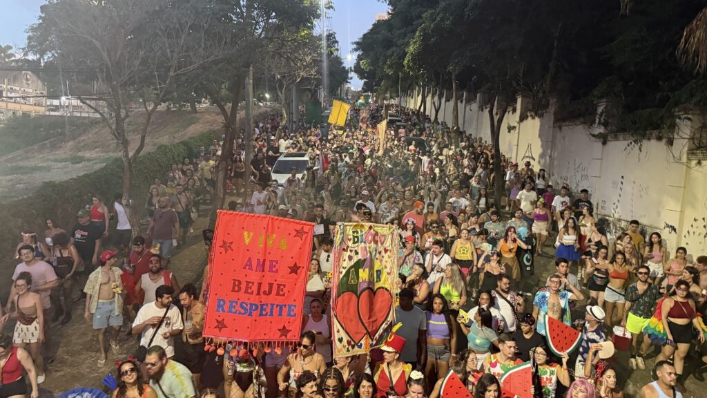 A imagem mostra uma grande multidão participando de um bloco de Carnaval nas ruas do Brasil. A foto foi tirada de um ângulo elevado, revelando uma rua estreita completamente tomada por pessoas que celebram animadamente. Muitos estão vestidos com roupas coloridas e fantasias típicas de Carnaval, algumas com acessórios divertidos como leques em forma de melancia. No centro da imagem, destacam-se dois grandes estandartes coloridos. Um deles é vermelho com a frase "VIVA AME BEIJE RESPEITE" escrita em letras grandes e coloridas, transmitindo uma mensagem de amor e respeito. O outro estandarte é mais decorativo, com detalhes em corações e franjas, carregando a inscrição "BLOCO DO AMOR". A atmosfera é festiva e vibrante, com pessoas sorrindo, dançando e celebrando juntas, refletindo a energia contagiante típica dos blocos de Carnaval brasileiros. Ao fundo, árvores e paredes laterais delimitam a rua, enquanto a luz do entardecer suaviza o cenário, criando um clima acolhedor e alegre.