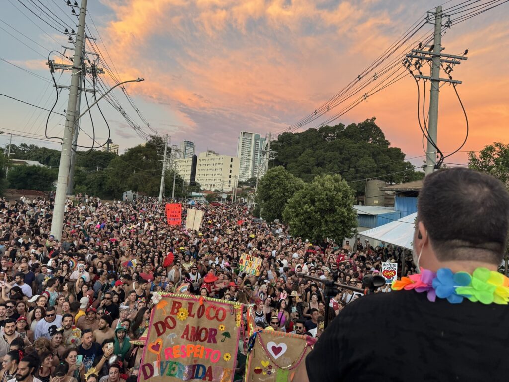 A imagem mostra uma grande festa de rua, típica dos blocos de Carnaval no Brasil, vista de um ponto elevado atrás de um participante. O céu ao fundo está tingido de tons vibrantes de laranja e rosa, indicando o pôr do sol. A multidão é enorme, composta por milhares de pessoas que vestem roupas coloridas, fantasias e adereços como colares de flores. No primeiro plano, vê-se a nuca de um homem com um colar de flores coloridas, olhando para a multidão que se estende até o horizonte, onde aparecem prédios altos e árvores verdes. Há faixas e cartazes pendurados, com mensagens que celebram o amor, o respeito e a diversidade, como “BLOCO DO AMOR RESPEITO DIVERSIDADE”, “VIVA AME BEIJE RESPEITE”, “AMOR É A ÚNICA RELIGIÃO” e “RESPEITO É REGRA”. A atmosfera é festiva, alegre e inclusiva, destacando valores de união e aceitação entre os participantes. A iluminação natural do pôr do sol cria um clima acolhedor e vibrante para o evento.