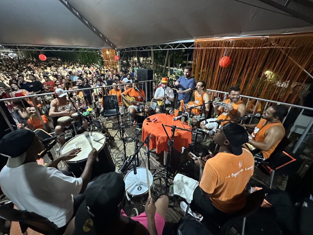 A imagem mostra um grupo musical de samba em uma apresentação ao vivo, realizada sob uma tenda branca à noite. O grupo é composto por cerca de oito homens, todos vestidos com camisetas ou regatas laranja com um logotipo, sentados em círculo ao redor de uma mesa redonda coberta por um pano laranja, onde há alguns copos e microfones. Eles tocam instrumentos típicos do samba, como surdo, pandeiros, cavaquinho, violão e repique de mão. À frente, um músico de costas, vestido com uma camisa branca e boné preto, toca um grande tambor. A apresentação ocorre em um espaço aberto, com uma grande plateia ao fundo, composta por muitas pessoas que assistem do outro lado de uma grade metálica. A iluminação é forte e artificial, destacando os músicos e criando uma atmosfera festiva. Há também uma decoração com franjas laranja penduradas, reforçando o clima de evento cultural ou festa popular. No uniforme dos músicos, é possível ler parcialmente o nome "Bagaço da Laranja", que provavelmente é o nome do grupo. A imagem transmite a energia e a alegria típicas de uma roda de samba, com músicos concentrados e público animado.