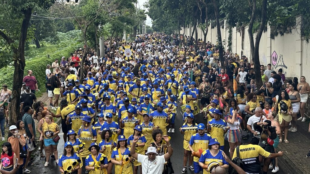 A imagem mostra um grande desfile de rua, possivelmente um bloco carnavalesco, com uma bateria (grupo de percussionistas) vestindo uniformes amarelos e azuis, cores que lembram o serviço postal brasileiro (Correios). Eles estão marchando por uma rua arborizada, cercados por uma multidão de espectadores que acompanham o evento. O clima parece nublado, com luz difusa. Na frente da bateria, um homem de camisa branca e chapéu levanta os braços, provavelmente conduzindo o grupo. Os músicos tocam diversos instrumentos de percussão típicos do samba, como surdos e chocalhos. De um lado da rua há árvores verdes e do outro uma parede branca com grafites. Ao fundo, banners indicam nomes como "Gil Coordenador" e "Norberto Coordenador de Bateria". A foto é tirada de um ponto alto, mostrando a extensão da festa e o grande número de participantes e espectadores, transmitindo uma atmosfera animada e festiva.