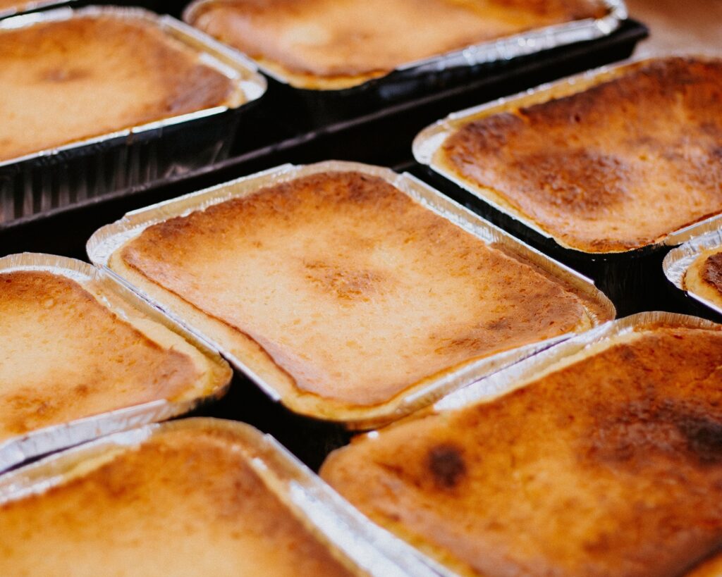 A imagem mostra uma fileira de tortinhas assadas, provavelmente tortinhas de milho, dispostas em bandejas retangulares de alumínio descartável. O foco está nas tortinhas centrais, que têm uma superfície dourada e levemente caramelizada, com uma textura lisa e opaca. As bordas das tortinhas parecem crocantes e levemente tostadas, conferindo um aspecto artesanal e apetitoso. A iluminação suave destaca as cores quentes do alimento, como tons de dourado, bege e âmbar, enquanto as bandejas metálicas refletem a luz, criando um contraste interessante. A profundidade de campo é rasa, deixando as tortinhas mais próximas e mais distantes um pouco desfocadas, o que direciona a atenção para o centro da imagem. A foto transmite uma sensação de frescor e cuidado na preparação das tortinhas.