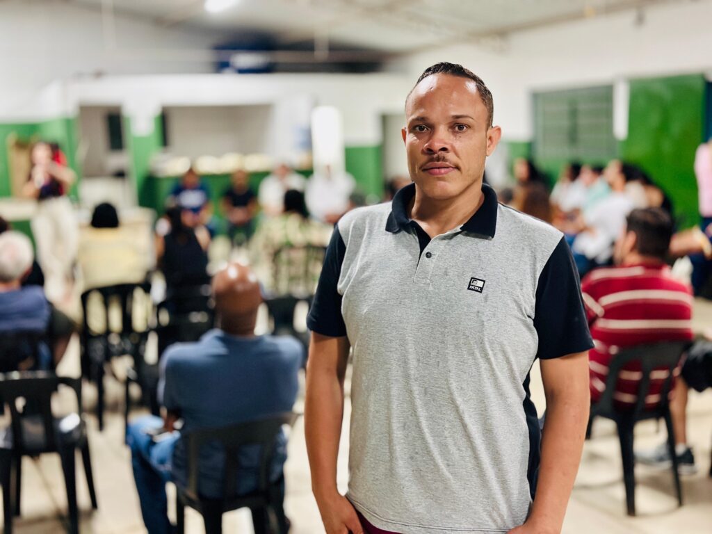 A imagem mostra um homem negro de pele clara, com bigode fino, olhando diretamente para a câmera com uma expressão neutra e calma. Ele veste uma camisa polo cinza com detalhes pretos e um pequeno logo no peito que diz "FATAL". O fundo está desfocado, revelando um ambiente interno amplo, possivelmente uma sala de eventos ou reunião, com várias pessoas sentadas em cadeiras pretas de plástico, sugerindo que estão participando de um encontro ou palestra. As paredes são brancas com detalhes verdes e a iluminação é clara e uniforme. O foco da imagem está no homem, destacando-o em meio ao cenário movimentado ao fundo.