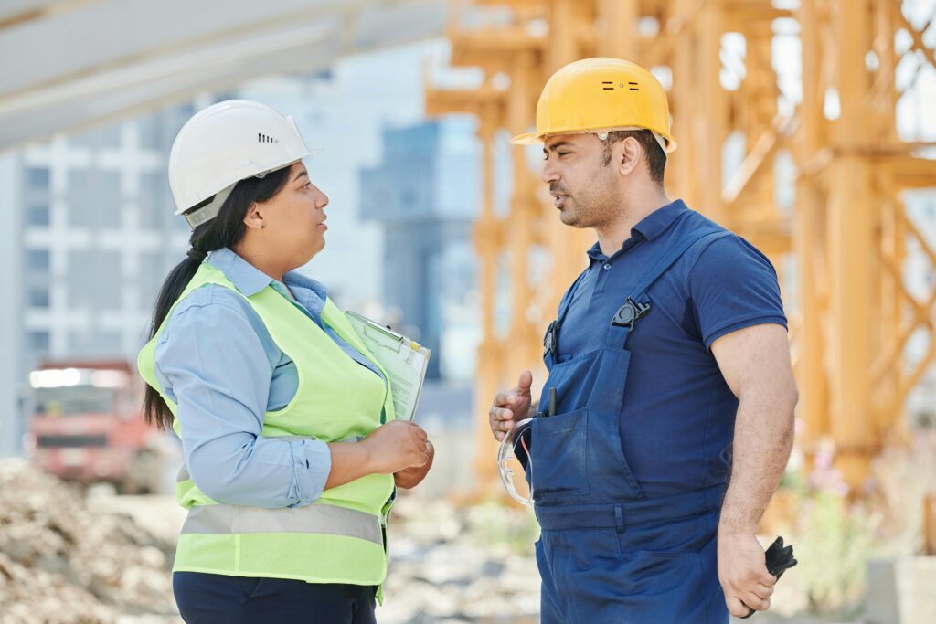 A imagem mostra dois trabalhadores da construção civil conversando em um canteiro de obras. À esquerda, há uma mulher usando capacete branco, uma camisa azul clara e um colete refletivo verde neon. Ela segura uma prancheta com papéis, parecendo atenta à conversa. À direita, um homem usa capacete amarelo, camiseta azul escura e macacão azul. Ele está gesticulando com a mão direita enquanto fala. Ao fundo, é possível ver estruturas de construção, incluindo um guindaste amarelo e um caminhão vermelho, indicando um ambiente ativo de obra. A iluminação é natural, sugerindo que a foto foi tirada durante o dia.