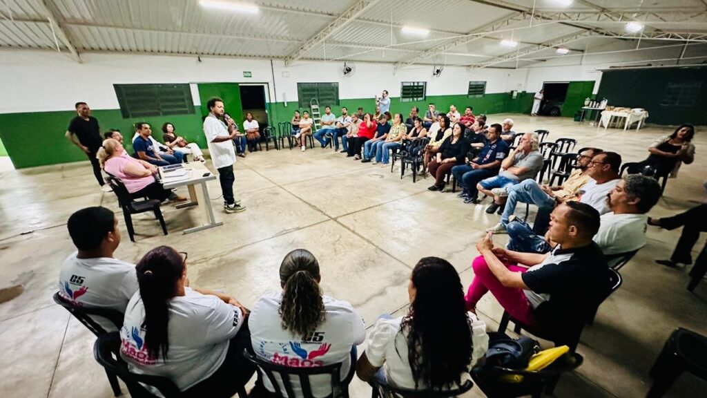 A imagem mostra uma reunião comunitária realizada em um salão amplo e simples. As pessoas estão sentadas em círculo, criando um ambiente de diálogo e participação igualitária. No centro da imagem, um homem está em pé, falando ao microfone, provavelmente liderando ou apresentando algo para o grupo. O salão tem paredes brancas com uma faixa verde, teto com estrutura metálica e iluminação fluorescente. Várias pessoas vestem camisetas brancas com um logo que mostra duas mãos segurando um coração, indicando que pode ser um grupo ou organização chamada "Mãos". Ao fundo, há uma mesa com alimentos e bebidas, sugerindo que o encontro pode ser prolongado ou informal em algum momento. A atmosfera geral é de uma reunião comunitária, onde as pessoas estão envolvidas e atentas ao que está sendo discutido.