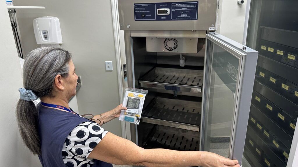 A imagem mostra uma mulher de cabelos grisalhos presos em um rabo de cavalo, vestindo um colete azul sobre uma blusa preta com padrão branco. Ela está em um ambiente clínico ou de laboratório, abrindo a porta de vidro de uma câmara conservadora metálica, que parece ser utilizada para armazenar vacinas ou medicamentos. Na mão esquerda, ela segura um medidor digital de temperatura, protegido por um saco plástico transparente. A câmara possui prateleiras metálicas perfuradas e um painel de controle na parte superior com um visor digital e instruções em português. Ao lado da câmara, há outro equipamento com várias gavetas rotuladas com nomes de vacinas ou doenças, como "ROTAVIRUS", "PNEUMO 10" e "DENGUE". O ambiente é bem iluminado e organizado, sugerindo um local dedicado à conservação adequada de produtos de saúde.