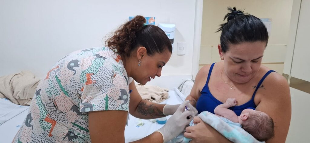 A imagem mostra uma cena em um ambiente clínico ou hospitalar, onde uma profissional de saúde está atendendo um recém-nascido que está nos braços da mãe. A profissional, à esquerda, veste um uniforme com estampas de animais (elefantes, girafas e zebras), usa luvas brancas e tem um cabelo castanho cacheado preso. Ela está concentrada no bebê, realizando algum procedimento ou exame. A mãe, à direita, veste uma regata azul e observa o bebê com um olhar calmo e atento, segurando-o com cuidado, envolto em um cobertor claro. O ambiente é claro e limpo, com paredes brancas e uma cama hospitalar ao fundo, além de um dispensador de sabonete ou álcool em gel na parede. A imagem transmite um momento de cuidado e atenção entre a profissional de saúde, a mãe e o recém-nascido.