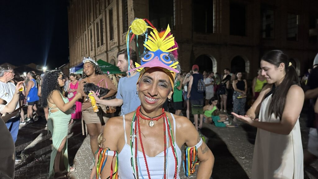 A imagem mostra uma mulher sorridente e alegre, vestida com uma fantasia vibrante e colorida que inclui uma grande peça de cabeça decorada com penas nas cores amarela, azul, rosa e vermelha, formando o formato de um pássaro. Ela usa vários colares de contas coloridas e um pingente de búzios, além de pulseiras com fitas coloridas nos braços. Ao fundo, há várias pessoas, algumas também vestidas de forma festiva, e um prédio antigo de tijolos com janelas arqueadas, sugerindo que o evento ocorre ao ar livre, à noite. A iluminação artificial destaca a mulher no centro da imagem, criando um contraste com o fundo mais escuro. A cena parece ser de um carnaval ou festa popular, transmitindo uma atmosfera de celebração e alegria.