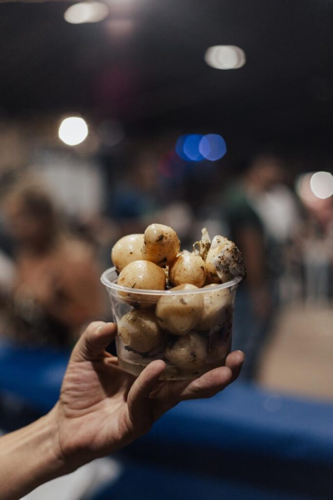 A imagem mostra uma mão segurando um pequeno recipiente transparente cheio de pequenas bolinhas claras, que parecem ser cebolinhas em conserva ou pequenos legumes em molho. O recipiente está em destaque, com foco nítido, enquanto o fundo está desfocado, apresentando várias luzes em formato de bokeh, sugerindo que a foto foi tirada em um ambiente interno à noite, possivelmente em um mercado ou restaurante. A mão é clara e segura o recipiente de forma firme, e a iluminação destaca os detalhes do alimento, realçando sua textura e cor. O fundo desfocado mostra algumas pessoas e estruturas, mas sem detalhes definidos, mantendo a atenção no recipiente de comida.