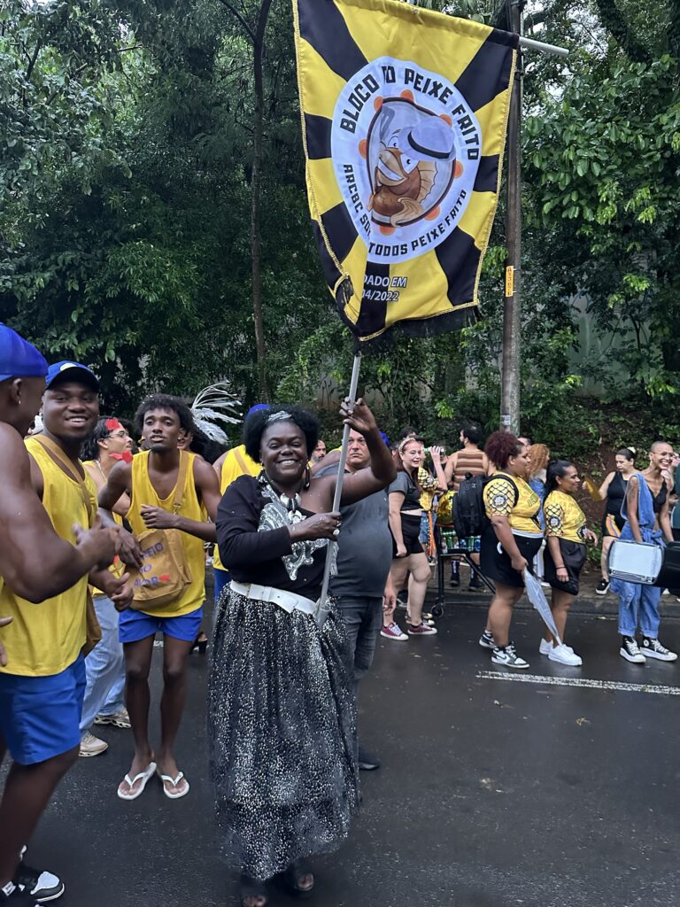 A imagem mostra um grupo de pessoas participando de um bloco de carnaval chamado "Bloco do Peixe Frito". No centro da foto, uma mulher sorri segurando uma bandeira amarela e preta com o desenho de um peixe, que é o mascote do bloco. Ela veste uma saia prateada brilhante e uma blusa preta, transmitindo alegria e energia. Ao redor dela, há outros foliões usando camisetas amarelas e shorts azuis, também sorrindo e caminhando pela rua molhada, indicando que pode ter chovido recentemente. O ambiente é ao ar livre, com árvores verdes ao fundo, sugerindo que o bloco está acontecendo em uma área urbana com bastante vegetação. A bandeira traz os dizeres "Bloco do Peixe Frito", "ArcBC Sou Todos Peixe Frito" e "Fundado em 04/2022", indicando que o grupo é recente. Um dos homens carrega uma bolsa preta com a frase "Feio do Amor". A atmosfera é festiva e animada, típica dos blocos de carnaval brasileiros.