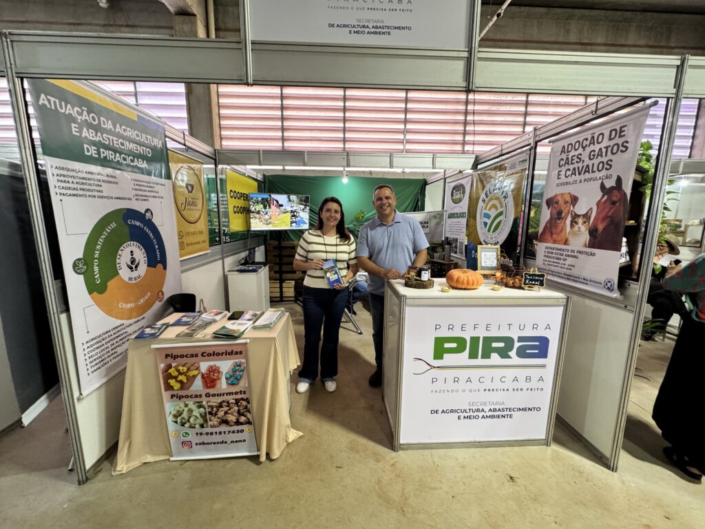A imagem mostra um estande promocional da Prefeitura de Piracicaba, mais especificamente da Secretaria de Agricultura, Abastecimento e Meio Ambiente. No estande, duas pessoas — uma mulher e um homem — estão sorrindo para a câmera, aparentando estar atendendo o público. O espaço está dividido em duas partes principais. À esquerda, há uma mesa coberta com uma toalha bege, sobre a qual estão expostos folhetos e materiais informativos, além de uma placa que anuncia "Pipocas Coloridas" e "Pipocas Gourmets", com um contato via WhatsApp e Instagram. Atrás dessa mesa, há um banner grande com o título "Atuação da Agricultura e Abastecimento de Piracicaba", listando várias ações e projetos da secretaria, como adequação ambiental rural, conservação de estradas, agricultura urbana, turismo rural, entre outros. À direita, há um balcão branco com a logomarca da Prefeitura de Piracicaba e o nome da secretaria. Sobre este balcão, estão expostos alguns objetos, incluindo uma abóbora, potes e uma pequena lousa com a inscrição "Turismo Rural". Também há um banner que promove a adoção de cães, gatos e cavalos, com informações de contato do Departamento de Proteção e Bem-Estar Animal. Ao fundo, uma televisão exibe imagens externas, possivelmente relacionadas às atividades rurais ou ambientais promovidas pela secretaria. A iluminação do local é clara e uniforme, sugerindo que o evento ocorre em ambiente fechado, como uma feira ou exposição pública. O estande passa uma imagem organizada, profissional e voltada para a divulgação de ações públicas ligadas ao meio ambiente, agricultura e bem-estar animal.