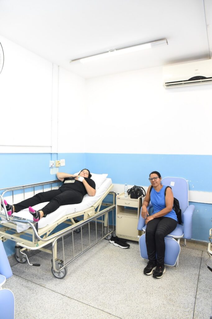 A imagem mostra uma cena em um ambiente hospitalar ou clínico. À esquerda, há uma mulher deitada em uma cama hospitalar com grades metálicas, vestindo uma camiseta preta e calça preta. Ela está com os olhos fechados, aparentando estar descansando, e tem uma atadura branca no braço esquerdo. Ela usa meia preta com detalhes em rosa. Ao lado da cama, há um pequeno armário de cabeceira bege, sobre o qual estão uma garrafa de água e uma bolsa preta. À direita, uma segunda mulher está sentada em uma poltrona médica azul claro, usando óculos, uma blusa azul sem mangas e calça preta. Ela olha para a câmera com uma expressão neutra. O ambiente tem paredes brancas com uma faixa azul na parte inferior, iluminação fluorescente no teto e um aparelho de ar-condicionado instalado na parede. O chão é revestido com um material cinza claro com manchas. A atmosfera parece tranquila e clínica.