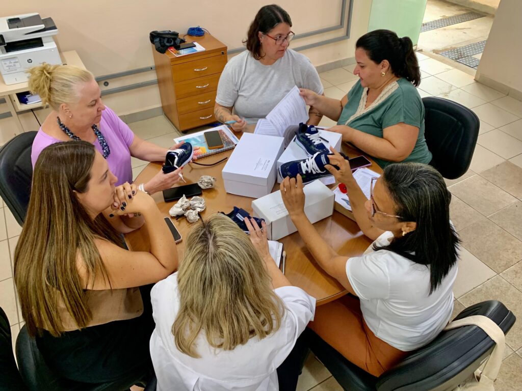 A imagem mostra seis mulheres sentadas ao redor de uma mesa redonda de madeira, em um ambiente interno que parece ser um escritório ou sala administrativa. Elas estão analisando vários pares de tênis azul-marinho com cadarços brancos e solado branco, que estão sobre a mesa, junto com algumas caixas brancas, papéis, celulares e pedaços de papel amassados. As mulheres estão concentradas, algumas segurando os tênis, outras olhando documentos. O local tem iluminação clara, provavelmente de luzes fluorescentes no teto. Ao fundo, há um armário de madeira e uma impressora HP sobre uma mesa. O chão é de azulejos claros. A cena transmite uma sensação de trabalho colaborativo, possivelmente uma reunião para controle de qualidade ou organização de produtos. A foto foi tirada de cima, mostrando a mesa e as pessoas em volta.