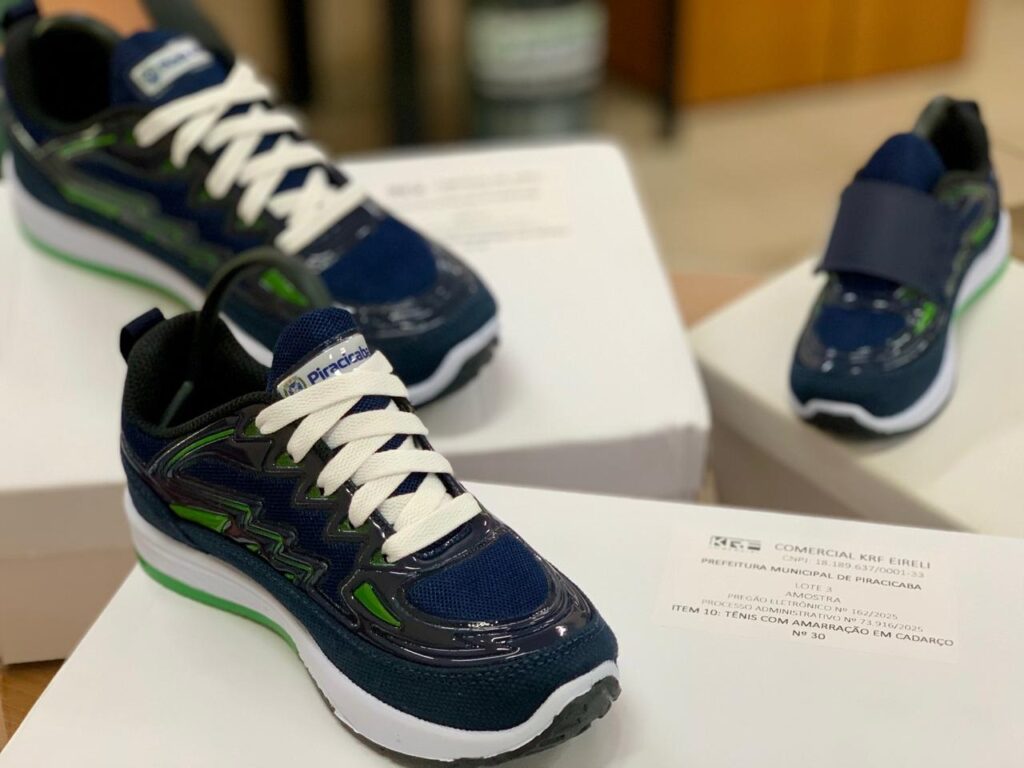 A imagem mostra um close de três pares de tênis esportivos de cor azul escuro, posicionados sobre caixas brancas. O tênis em destaque, à frente, possui cadarços brancos grossos, uma parte superior em mesh azul marinho com detalhes brilhantes e detalhes em verde limão. Ao fundo, à esquerda, há outro tênis parecido com o da frente, e à direita, um modelo diferente com fechamento em velcro em vez de cadarço. A sola dos tênis é branca com uma faixa verde próxima ao calcanhar. No canto inferior direito da imagem, aparece uma etiqueta branca com informações em português, indicando que o produto é uma amostra para a Prefeitura Municipal de Piracicaba, mencionando também um pregão eletrônico e um processo administrativo. O ambiente ao fundo está desfocado, sugerindo um local interno.
