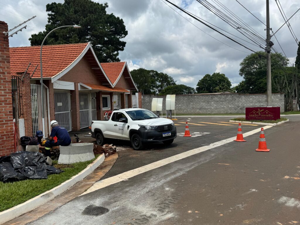 A imagem mostra uma cena de manutenção na entrada de um condomínio residencial chamado "Moinho Vermelho Residencial". Dois trabalhadores estão realizando reparos próximos a uma construção de tijolos com telhado de cerâmica vermelha. Um dos trabalhadores usa capacete branco e uniforme azul, sentado próximo a um anel de concreto, enquanto o outro, com boné azul, está agachado ao lado dele. Ao lado, há uma caminhonete branca da marca Fiat, com a inscrição "SANE SOLUTIONS" e "Semae" nas portas, além da placa de identificação "TEL9G54". No chão, quatro cones de sinalização laranja delimitam a área de trabalho. Ao fundo, há um muro de blocos cinza, árvores verdes e o céu está nublado. Linhas de energia elétrica cruzam a parte superior direita da imagem. A iluminação é suave, típica de um dia nublado, e a foto foi tirada de um ângulo ligeiramente elevado, mostrando a relação entre o serviço de manutenção e a entrada do condomínio.