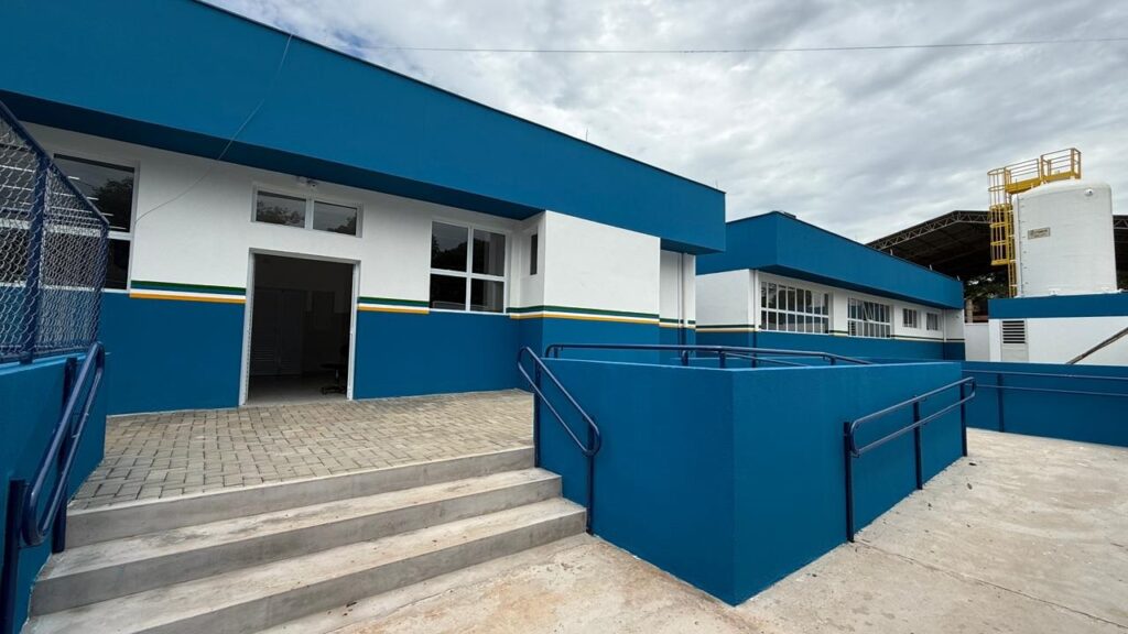 A imagem mostra um edifício moderno de um único pavimento sob um céu nublado. A construção apresenta uma combinação de cores vibrantes, com as paredes pintadas em azul e branco, separadas por faixas horizontais finas nas cores amarela e verde. Na fachada principal, há uma porta de entrada ampla, várias janelas com molduras brancas e uma rampa de acesso com corrimãos metálicos azuis, garantindo acessibilidade. O piso em frente ao prédio é feito de blocos de concreto intertravados, e há três degraus de concreto que levam até a entrada. À direita, observa-se uma outra parte do edifício, além de um grande tanque branco vertical e uma estrutura metálica amarela ao fundo. A fotografia foi tirada de um ângulo baixo, destacando as linhas retas e o acabamento limpo da construção. O ambiente parece ser uma instalação pública ou institucional, como uma escola ou centro de saúde, devido à arquitetura funcional e aos elementos de acessibilidade. Não há pessoas visíveis na imagem.