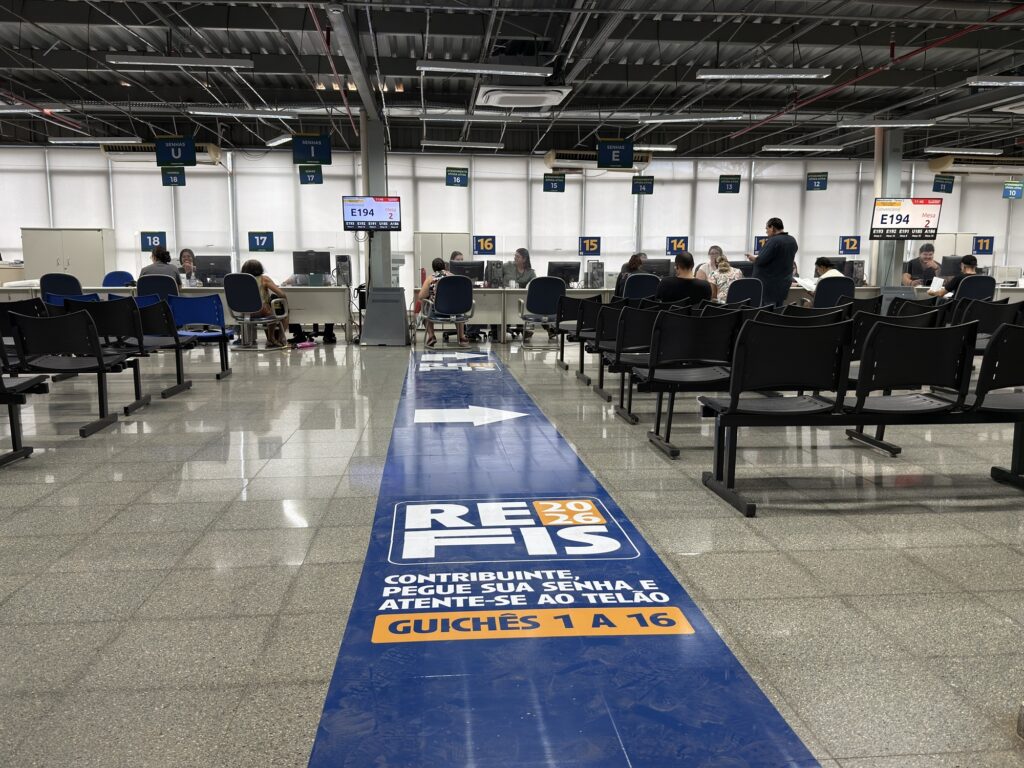 A imagem mostra uma sala ampla de atendimento público, provavelmente em um órgão governamental brasileiro, onde as pessoas aguardam para serem atendidas. No centro do espaço, há uma faixa azul no chão com o texto "REFIS 2023-2026", indicando um programa de refinanciamento de dívidas. À frente, há várias cadeiras pretas enfileiradas para os contribuintes sentarem enquanto esperam. Na parte de trás da sala, há vários guichês numerados de 1 a 16, onde funcionários atendem os cidadãos. Acima dos guichês, há um painel eletrônico que exibe senhas e o número do guichê para onde o próximo cidadão deve se dirigir. Também há placas com instruções em português, como “CONTRIBUINTE, PEGUE SUA SENHA E ATENTE-SE AO TELÃO”. O ambiente é iluminado por luz natural que entra pelas janelas, além de luz artificial fluorescente. O teto tem um design industrial com tubulações aparentes. O piso é de granito polido, refletindo a luz do ambiente. A cena transmite uma atmosfera organizada e funcional, típica de um local de atendimento ao público para questões fiscais ou administrativas.