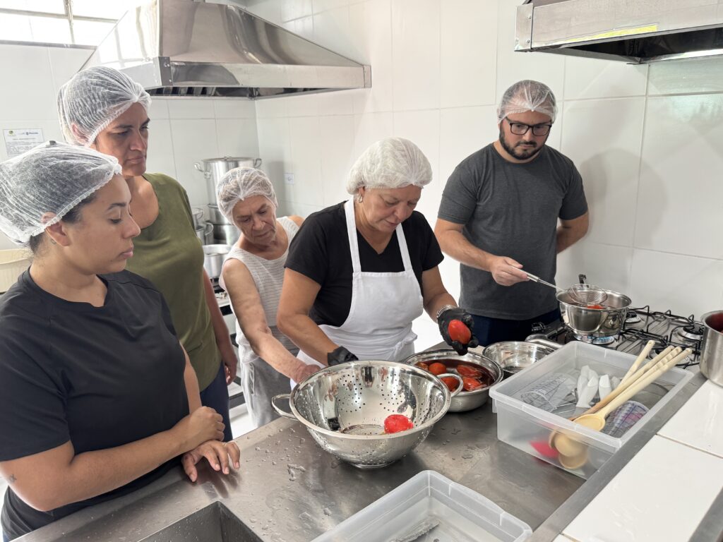 A imagem mostra um grupo de cinco pessoas em uma cozinha comercial, possivelmente em uma aula de culinária ou em um ambiente de cozinha comunitária. No centro, uma mulher usa avental branco, luvas pretas e está descascando ou manipulando tomates que parecem ter sido escaldados. Ao redor dela, outras três mulheres observam atentamente o processo; todas usam toucas para cabelo e roupas casuais. À direita, um homem de barba e óculos, também com touca, está usando uma escumadeira para retirar tomates de uma panela com água fervente sobre um fogão industrial. A cozinha é limpa, com paredes revestidas de azulejos brancos e superfícies de aço inoxidável. Acima do fogão, há um coifa grande de aço inox. Na bancada, há uma grande escorredora com tomates já descascados, uma tigela com água e tomates e um recipiente com utensílios de cozinha, como colheres de pau e copos medidores. A iluminação é clara e uniforme, criando um ambiente propício para o aprendizado ou trabalho em equipe. A palavra "CIÊNCIA" e o nome "Piracicaba" aparecem em um pequeno cartaz na parede ao fundo, sugerindo que a atividade pode estar relacionada a um projeto ou instituição local.