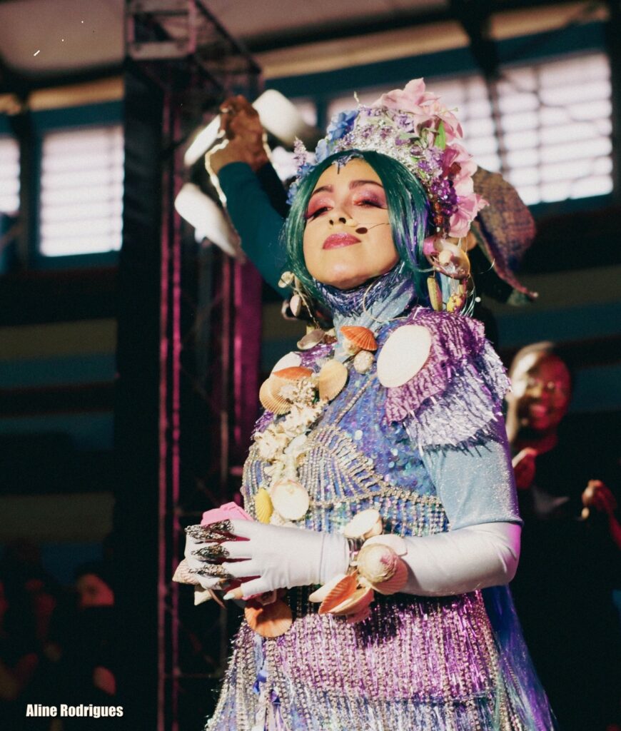 A imagem mostra uma pessoa em um palco, vestida com uma fantasia elaborada inspirada no tema do oceano. A fantasia é vibrante, combinando cores como azul-turquesa, roxo, prata e rosa, e é adornada com conchas marinhas de vários tipos e tamanhos. A pessoa usa uma peruca azul-turquesa e maquiagem rosa nos olhos, além de luvas brancas com unhas longas prateadas que lembram garras. A cabeça está decorada com uma coroa feita de flores e conchas. A iluminação do palco usa tons rosados e brancos, criando um efeito dramático, enquanto o fundo está desfocado, destacando o personagem principal. A expressão da pessoa é serena, olhando para cima, transmitindo uma aura mística ou de contemplação. O nome "Aline Rodrigues" aparece no canto inferior esquerdo, possivelmente indicando o nome do fotógrafo ou do artista. A cena sugere um espetáculo teatral, show de drag ou carnaval com tema marítimo ou de sereia.