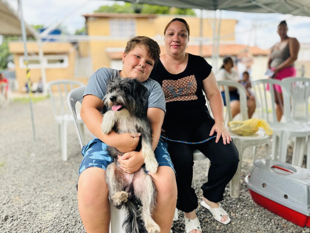 Um menino está sentado em uma cadeira plástica branca, com um cachorro preto e branco no colo. Ao lado dele, uma mulher — mãe do menino — está sentada e mantém a mão apoiada na perna do garoto, transmitindo proximidade e cuidado. Eles estão em um espaço externo coberto por uma tenda, com outras pessoas e cadeiras semelhantes ao fundo. O chão é de pedrinhas, e próximo à mulher há uma caixa de transporte para animais, vermelha e cinza.